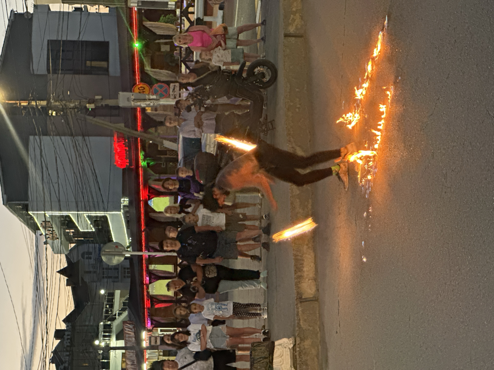 A fire performer is spinning a fiery prop on a street in Vityazevo, Russia. The performer, dressed in dark pants and a light-colored t-shirt, is creating a trail of fire on the asphalt. A crowd of onlookers, including men, women, and children, stands on the sidewalk behind a low concrete barrier, observing the performance. They are dressed in casual clothing. In the background, there are buildings with illuminated signs and a streetlamp. A black scooter is parked on the sidewalk to the right of the performer. The sky is a dusky orange and gray, indicating it is either dawn or dusk.