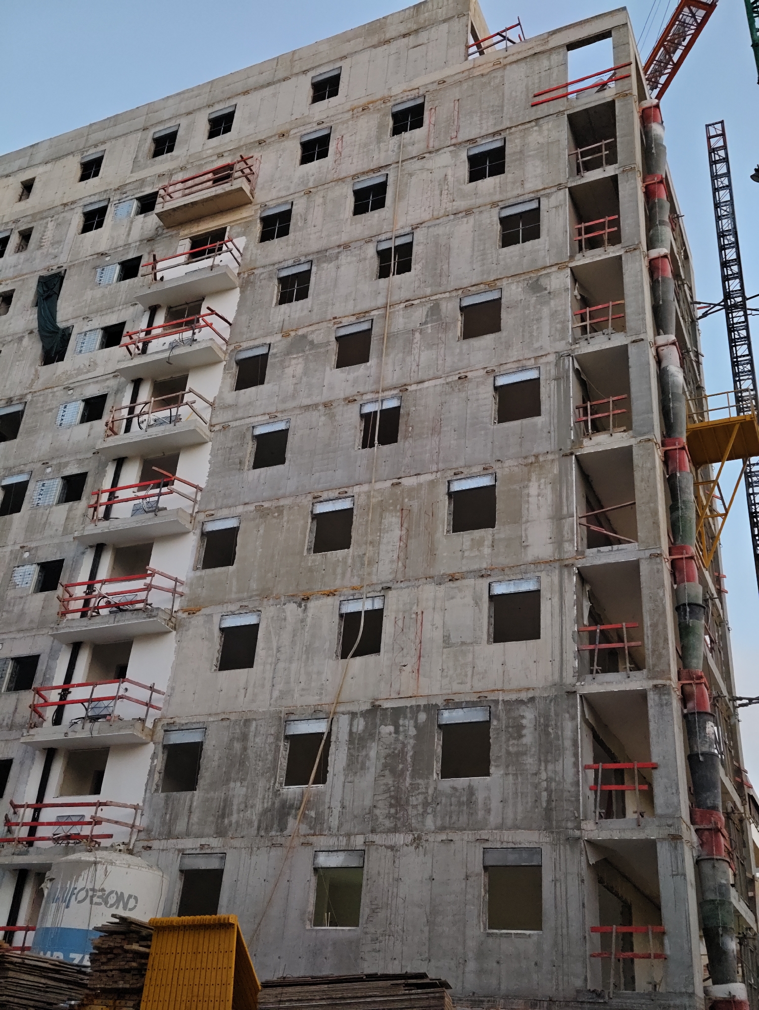 This image shows a partially constructed, multi-story concrete building in Petah Tikva, Israel. The building appears to be a residential or commercial complex, with multiple floors featuring balconies and numerous window openings, many of which are covered with a reflective material. Construction cranes are visible in the background, indicating ongoing work. There are no people clearly visible in the image, but construction materials such as wooden planks and yellow protective barriers are present at the base of the structure. The overall scene depicts a typical construction site in an urban environment.