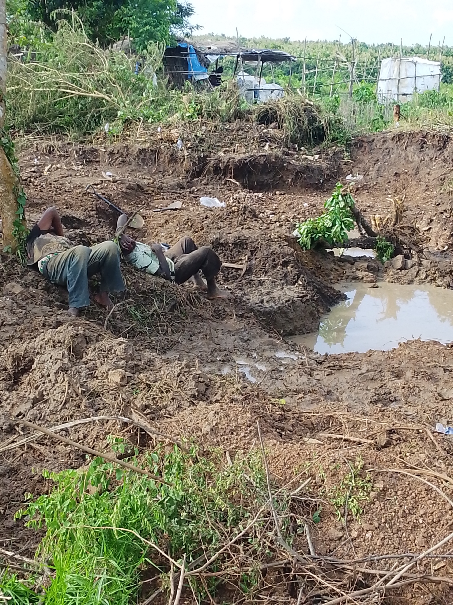 The image depicts an outdoor scene in Otulu, Nigeria, characterized by a muddy, debris-strewn environment, likely after a natural event such as a landslide or heavy rainfall. In the foreground, two individuals are lying down, appearing exhausted or possibly injured, amidst the mud and scattered vegetation. They are dressed in casual clothing. One person is lying on their back, propped up by their arm, while the other is lying on their side. Tools, such as what appears to be a shovel, are visible in the vicinity, suggesting manual labor or rescue efforts. In the background, there are makeshift structures and dense vegetation, indicating a rural or semi-urban area. The ground is uneven and appears to have been recently disturbed, with exposed soil and a small pool of water. The overall impression is one of hardship and recovery in a challenging environment.