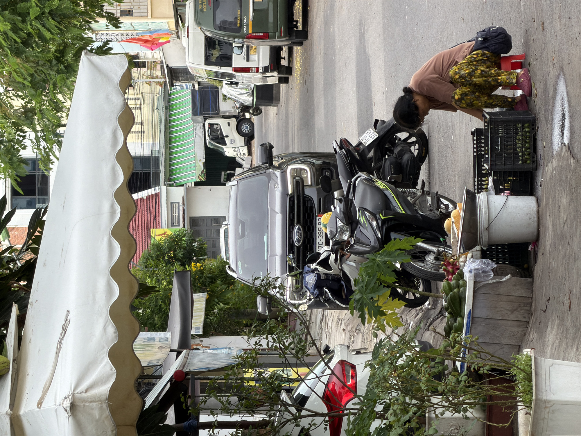 The image, taken in Nha Trang Ward, Vietnam, depicts a vibrant street scene with a person and various vehicles alongside market stalls. In the mid-ground on the right, a woman is seen bending over, seemingly arranging or inspecting produce in black plastic crates on the sidewalk. She is wearing a brown shirt, patterned yellow-green pants, and pink shoes, with a black backpack resting on a red plastic stool behind her. The crates contain green fruits, possibly limes or small gourds, and other fruits like a red dragon fruit and a bunch of green bananas are visible nearby, resting on a white bucket.
In the foreground on the left, a large white market awning with scalloped edges extends into the frame. Behind it, lush green foliage from trees and bushes lines the street. The street itself is busy with parked and moving vehicles. Several motorbikes, including black and grey models, are parked near the woman's stall. Further down the street, a dark grey Ford truck/van is visible, displaying "Ford" and a license plate "7C-395". Behind it, a white delivery truck and a green van with "KIM LONG" and a phone number "0919.331 883" are parked. Another white van with license plate "796 034" and text "XE TE HOTLI" (likely "XE TỐT HOTLINE") is also present. Buildings with different colored awnings (including green and white striped) line the street in the background. The scene is brightly lit, indicating daytime.