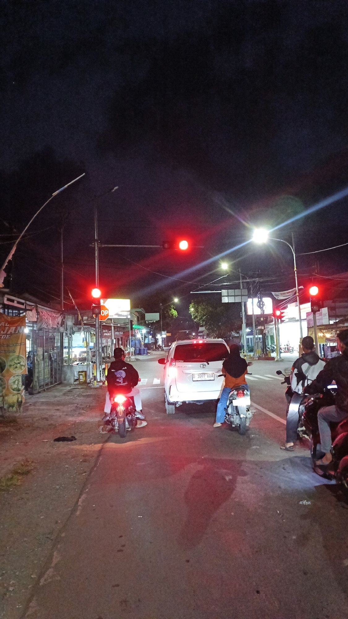 The image depicts a nighttime street scene in Desa Cijulang, Indonesia. The primary focus is an intersection with multiple vehicles and pedestrians under a red traffic light. In the foreground, two motorcycles are stopped, with riders facing away from the camera. The motorcycle on the left has a rider wearing a dark jacket with a graphic design. The motorcycle on the right has a rider wearing a hijab and blue pants. A white car is stopped between the two motorcycles, facing forward. To the right, another motorcycle is partially visible with two individuals on it. The street is paved and appears to be wet in places, reflecting the red light from the traffic signal. Buildings line the left side of the street, with signage visible, suggesting commercial establishments. Streetlights and overhead wires are also present. The sky is dark, indicating it is night. The overall atmosphere is that of a typical urban intersection at night, with traffic waiting for the light to change.