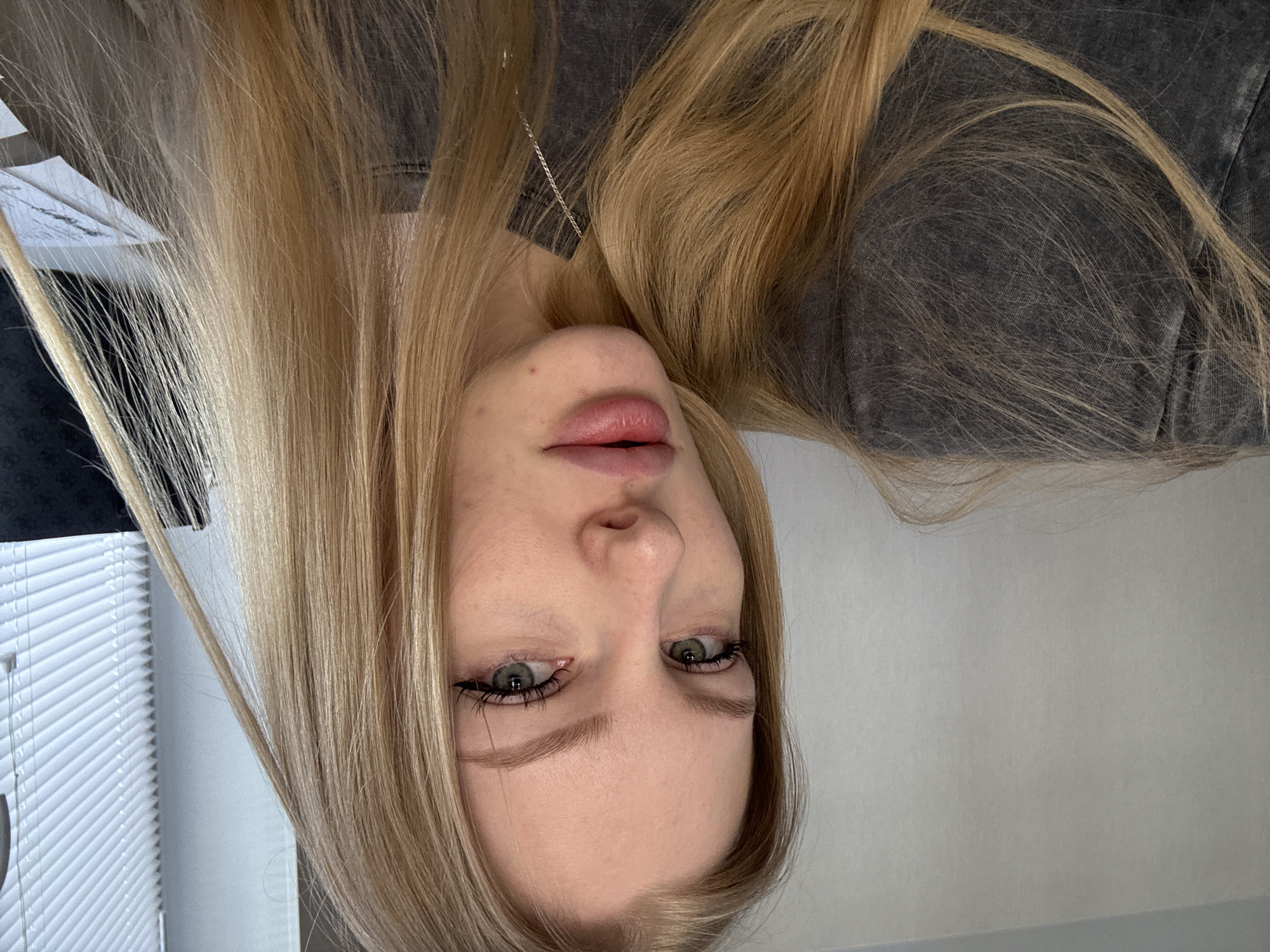 A blonde woman with light skin and green eyes is pictured in a close-up, upside-down shot. She has a gray denim shirt and a silver necklace. There is a set of white blinds in the background, and a white wall to the left. The image does not appear to contain information to confirm its origin is Perm, Russia.
