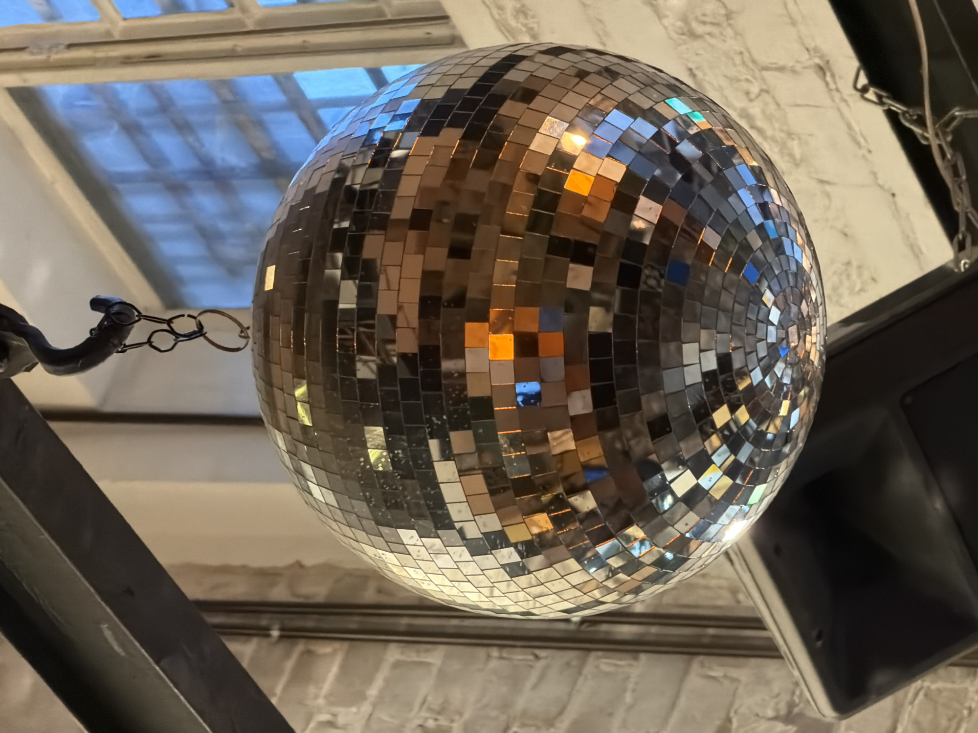 A disco ball hangs from a metal beam, its mirrored surface reflecting the light. The disco ball is covered in numerous small, square mirrors, creating a mosaic effect. In the background, a window with multiple panes allows light to filter in, illuminating a brick wall. A black speaker is positioned below the disco ball, and a chain hangs to the right. The scene is lit with warm, ambient light, creating a festive atmosphere. The location is confirmed to be in Tel Aviv, Israel.