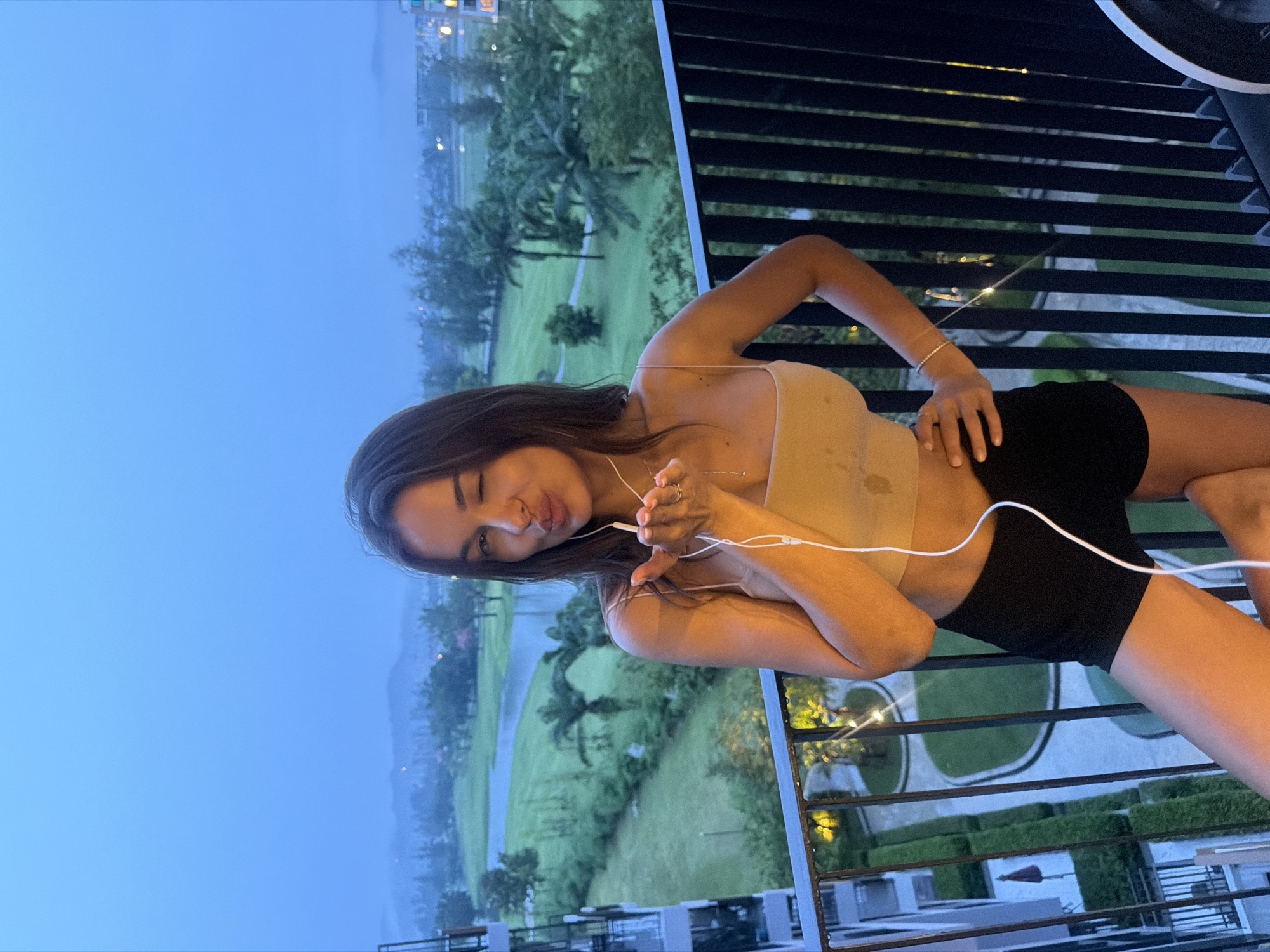 A woman poses on a balcony in Choeng Thale, Thailand, with a view of green landscapes and the evening sky. She wears a tan top and black shorts, holding white earphones. The balcony has black railings, and the background shows buildings, trees, and a golf course.