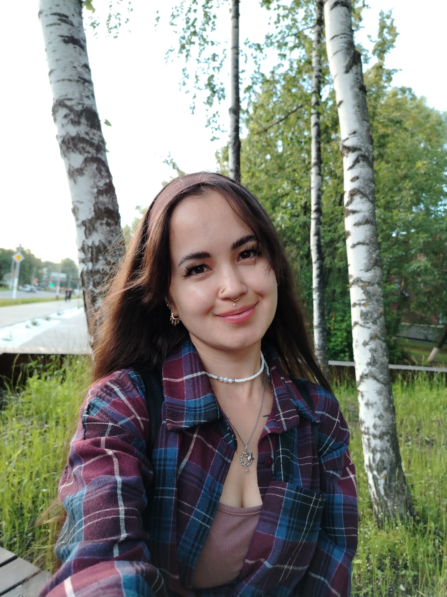 A young woman with long, dark brown hair and a nose ring smiles at the camera. She is wearing a maroon and blue plaid shirt over a pinkish-brown tank top, a white beaded necklace, and a silver pendant. A brown headband is on her head, and she has hoop earrings. The background is a park with several birch trees with white bark and green foliage. Lush green grass and a wooden bench are visible in the foreground. A street with cars and traffic lights can be seen in the distance, suggesting an urban park setting. The lighting indicates it is daytime, likely late afternoon, with soft sunlight filtering through the trees. The woman appears relaxed and happy. No visible text is present in the image. The location context is Chaikovsky, Russia.