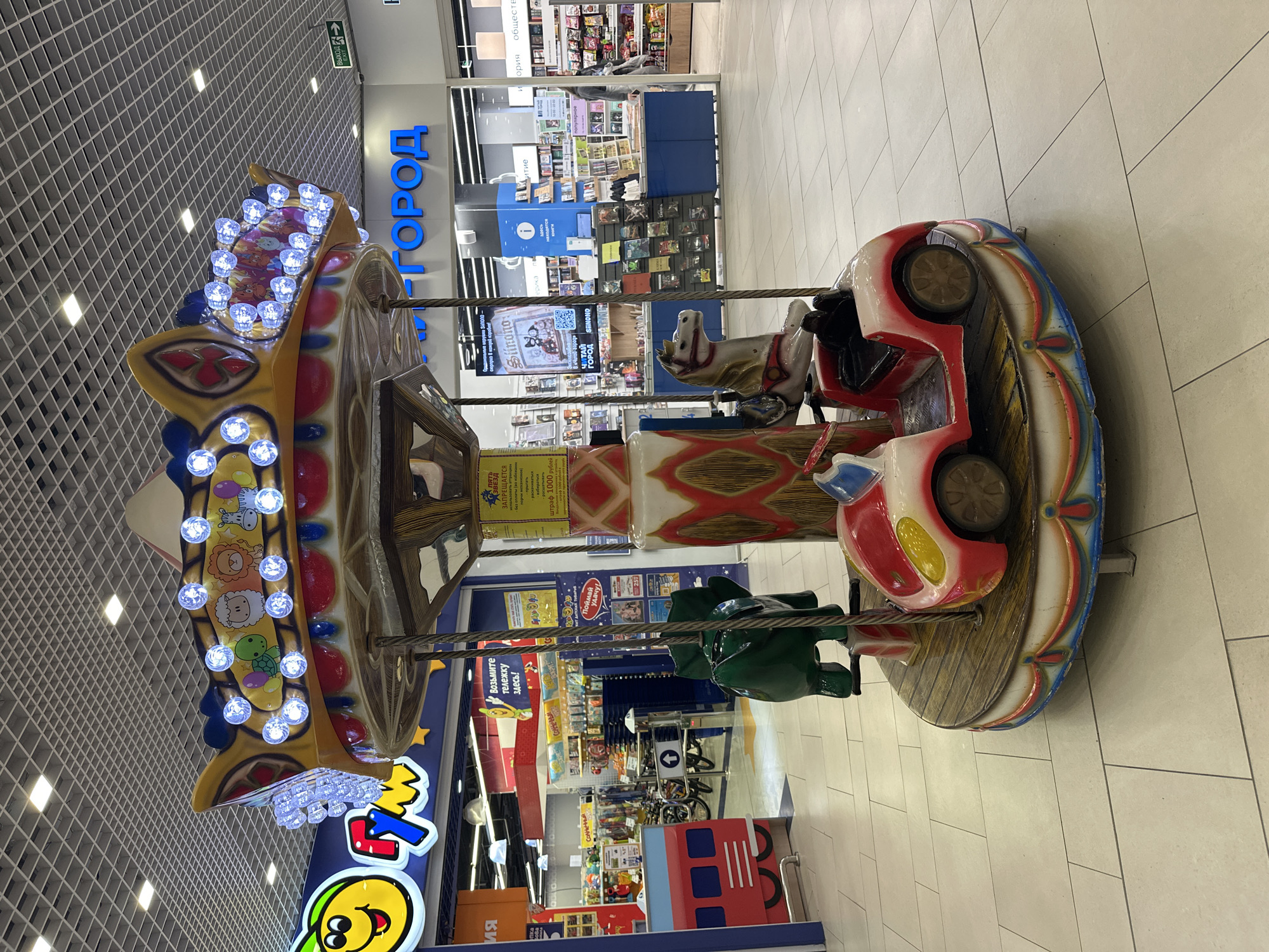 The image shows a brightly colored children's carousel located inside a shopping mall in Yekaterinburg, Russia. The carousel features ride options including a horse, car, and green alligator. The carousel is positioned between two retail stores: "Город" (Gorod), a bookstore, and a children's toy store. The floor is tiled. The ceiling is a white grid with embedded lighting fixtures. There are no people visible on the carousel.