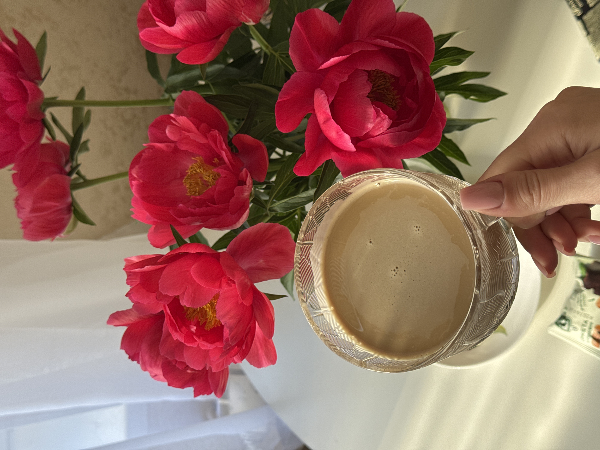 A close-up, eye-level shot shows a hand holding a glass cup of coffee with a frothy top. In the background, a bouquet of bright pink peonies is displayed in a vase. The scene is set on a white table. Sunlight casts a warm glow, creating shadows on the table and the white curtain in the background. The image is taken indoors, likely in a domestic setting, and the flowers and coffee suggest a moment of relaxation or a pleasant start to the day. There is no specific indication of the city of Chaikovsky, Russia, within the image itself.