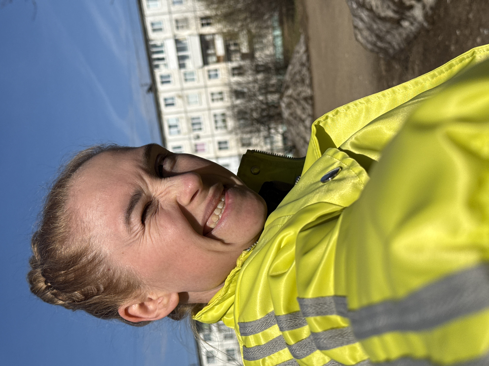 A selfie captures a fair-skinned individual smiling broadly with their eyes squinted against the bright sunlight. Their light brown hair is pulled back, and they are wearing a vibrant, almost neon, yellow jacket with horizontal grey reflective stripes on the sleeves and torso. The jacket appears to be made of a smooth, slightly shiny material.
The setting is outdoors on a sunny day with a clear blue sky visible in the upper left portion of the image. In the background, a large, light-colored multi-story residential building with numerous windows is visible, slightly out of focus. Below the building and to the right, the ground appears to be a mix of dirt and possibly melting snow or ice, suggesting a transitional season like early spring or late winter. The bright light and squinting eyes indicate a sunny time of day. The person appears joyful and happy.
The image is captured in Chaikovsky, Russia. There is no visible text in the image.