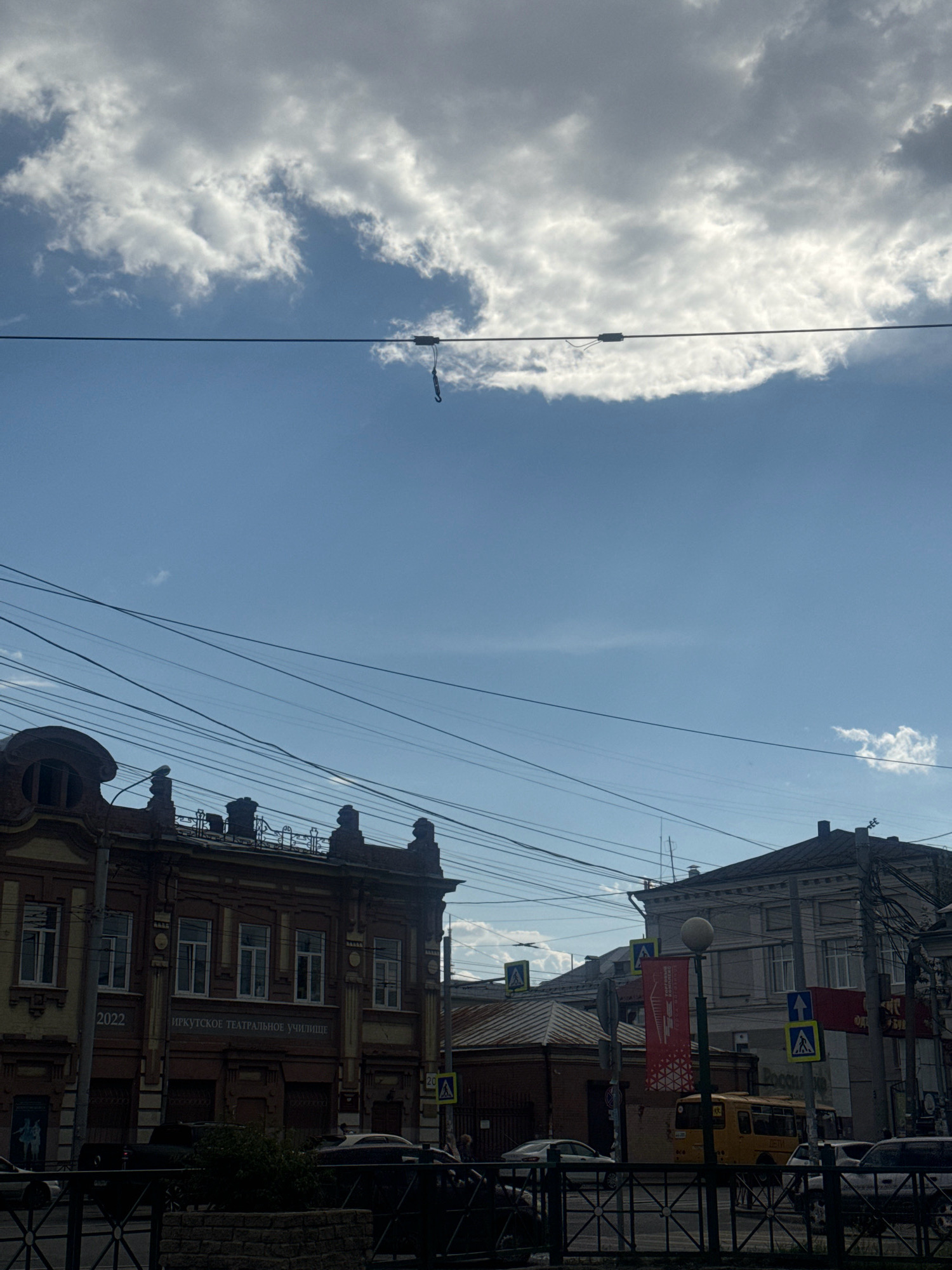 This is a medium shot, eye-level, outdoor photograph taken on a cloudy day, looking up at the sky and streetscape of Irkutsk, Russia. The prominent elements include a clear blue sky with scattered clouds, a network of overhead wires, and the upper facades of several buildings. A dark, hook-like object hangs from one of the wires, suspended in the middle of the frame.
On the left, a section of an ornate, brick building is visible. It features a rounded architectural element at the top left, several windows with white frames, and decorative brickwork. Text on the facade reads "2022" and "ИРКУТСКОЕ ТЕАТРАЛЬНОЕ УЧИЛИЩЕ" (Irkutsk Theater School).
To the right of this building, another building with a lighter facade and a sloped roof is partially visible. Further to the right, a gray building with multiple windows and a dark roofline dominates the right side of the frame. A yellow bus is parked on the street in front of this building. Several traffic signs, including pedestrian crossing signs and a directional sign, are visible. Red banners with geometric patterns hang from lampposts.
Below the buildings, a black metal fence is in the foreground, with a low brick wall behind it. The lower part of the image is dark, obscuring details of the street level. The overall impression is of an urban environment with historical architecture and active street life.