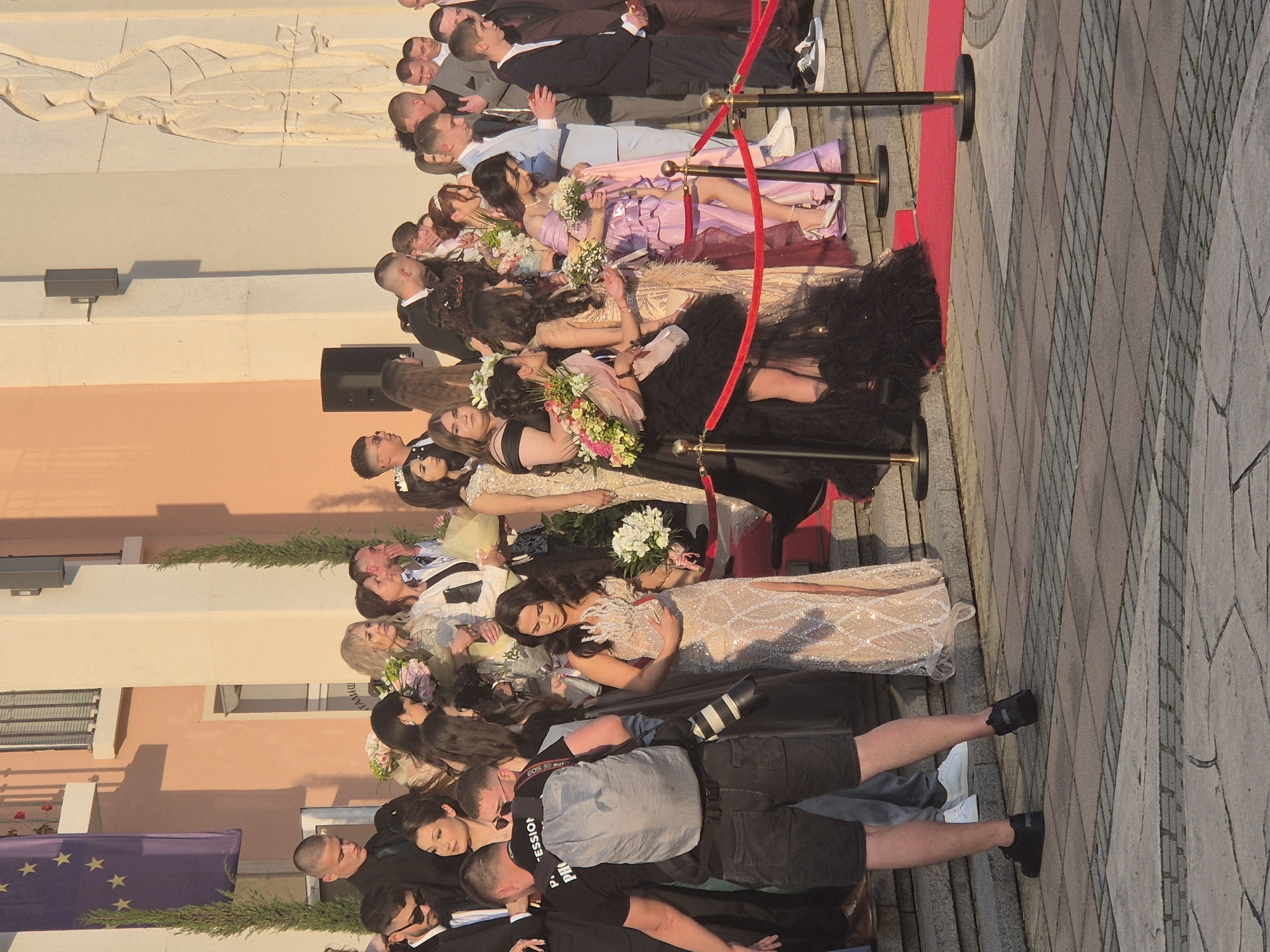 A group of people dressed in formal attire stand along a red carpet, lined by stanchions with a red rope. They are arranged on a grey stone tiled surface near a light-colored building. Some people are holding bouquets. A photographer is present, taking pictures with a professional camera. An EU flag is visible in the background. The event is located in Pavlikeni, Bulgaria.