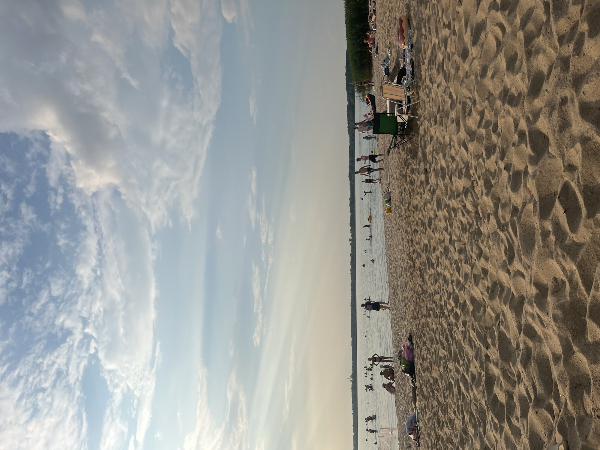 The image depicts a sandy beach adjacent to a body of water, likely a lake or river, under a partially cloudy sky. The beach is populated with numerous individuals engaged in various activities. In the foreground, the sand is textured with footprints and ripples. Towards the middle ground, people are seen wading and swimming in the water. Some individuals are sitting or lying on the sand, some on beach towels and blankets. Two beach chairs are set up on the sand. In the distance, a line of trees forms the horizon. The overall scene suggests a recreational setting on a warm day. The provided hints suggest the location is within the Загародскі сельскі Савет, Belarus.