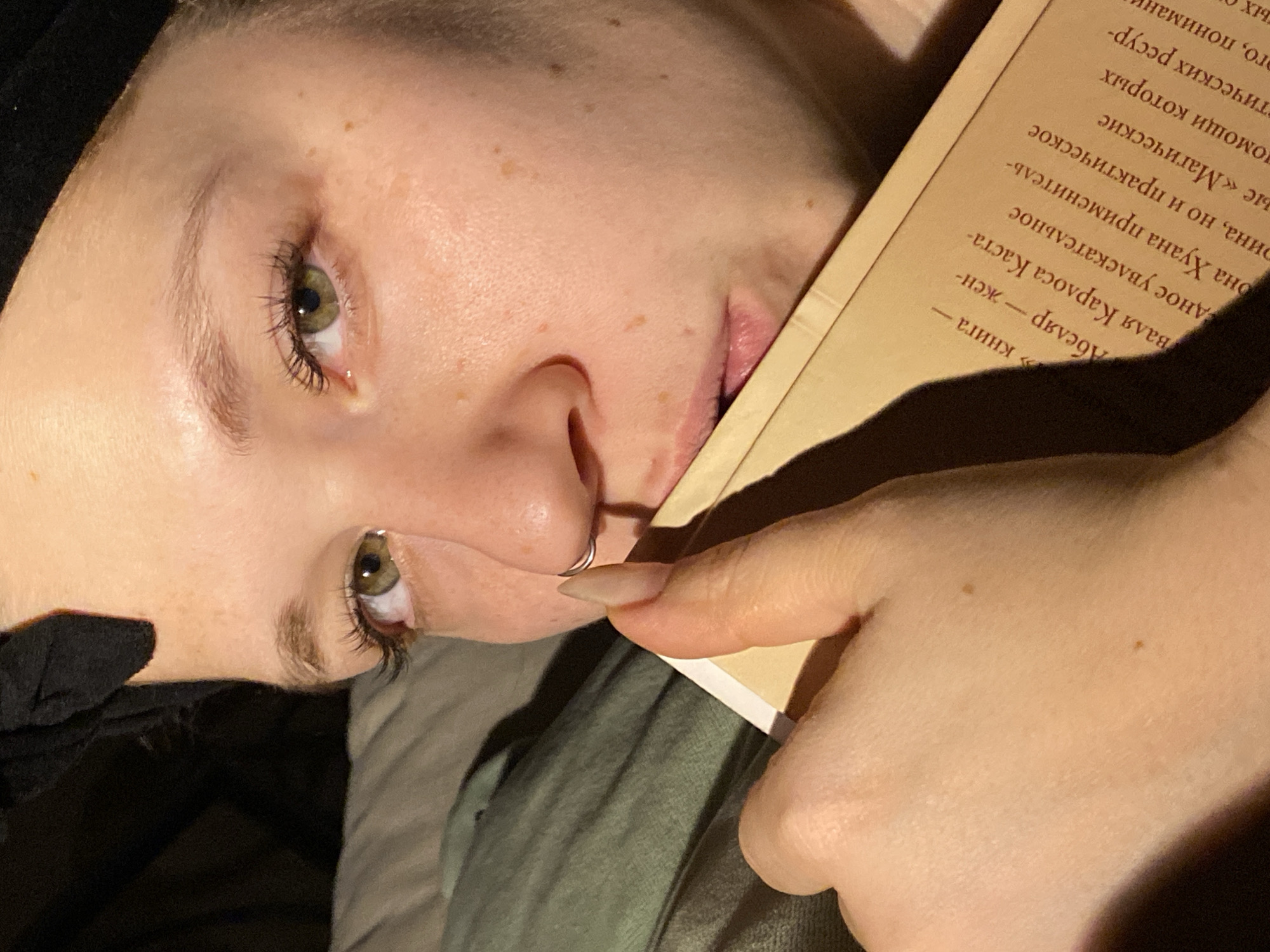 The image shows a close-up of a person's face as they are reading a book. The person is lying down, with the book propped up next to their face. The book's pages are visible and contain text in Russian, mentioning "Абеляр - жен-валя Карлоса Каста-". The person has fair skin, green eyes, and a nose ring. One hand is holding the book open. The background is dark and blurry. There is no indication of the city of Chaikovsky, Russia in the picture.