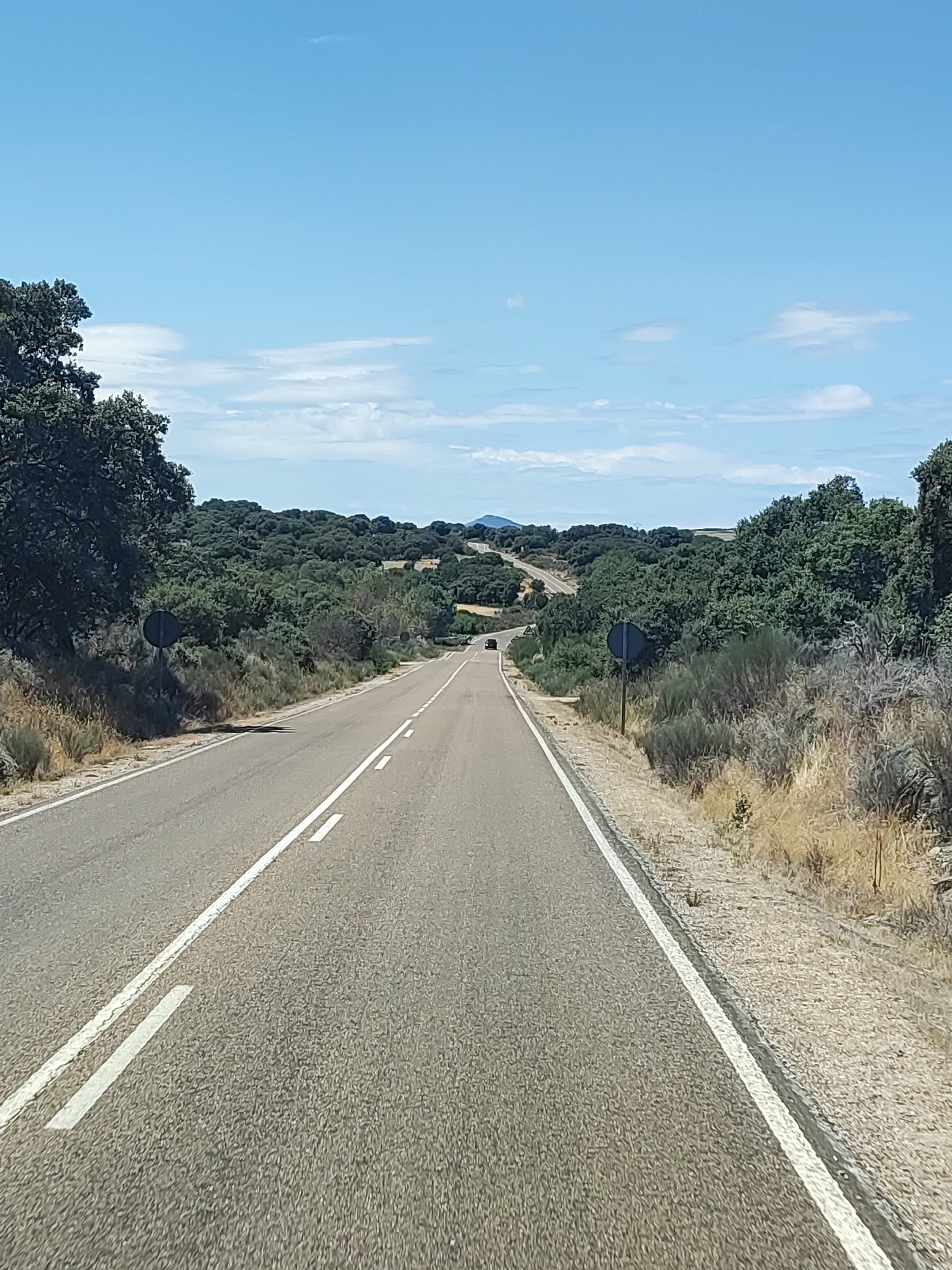 The image depicts a rural highway in the municipality of La Redonda, Spain, on a clear day. The road, paved with asphalt, features a solid white line on the right edge and a dashed white line separating two lanes. The road curves gently to the left as it recedes into the distance, with low green trees and bushes lining both sides. In the background, rolling hills covered with dense vegetation are visible under a bright blue sky with scattered white clouds. A dark-colored vehicle is present on the road in the distance, traveling in the same direction as the viewer. Two circular road signs are positioned along the right side of the road. The environment appears dry and sunny.