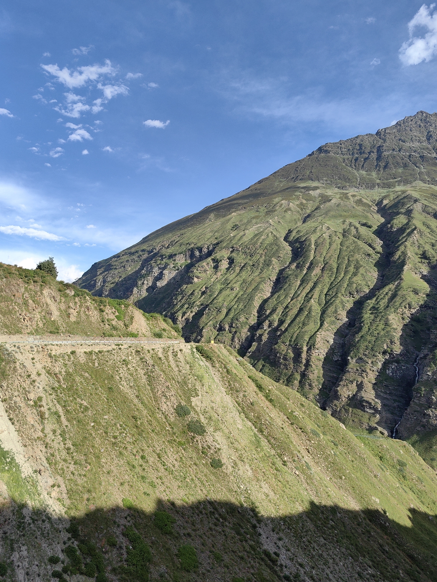 A view of a steep, green, mountainous landscape is captured under a bright blue sky with scattered white clouds. The mountain slopes are covered in a mix of grass and exposed rock, with visible ravines and shadows indicating uneven terrain. A narrow, paved road with a white guardrail winds along the side of a lower, grassy embankment in the foreground, suggesting it is a mountain pass or road through the area. Utility poles with wires are visible on the slopes. The overall scene conveys a sense of remote, natural beauty.