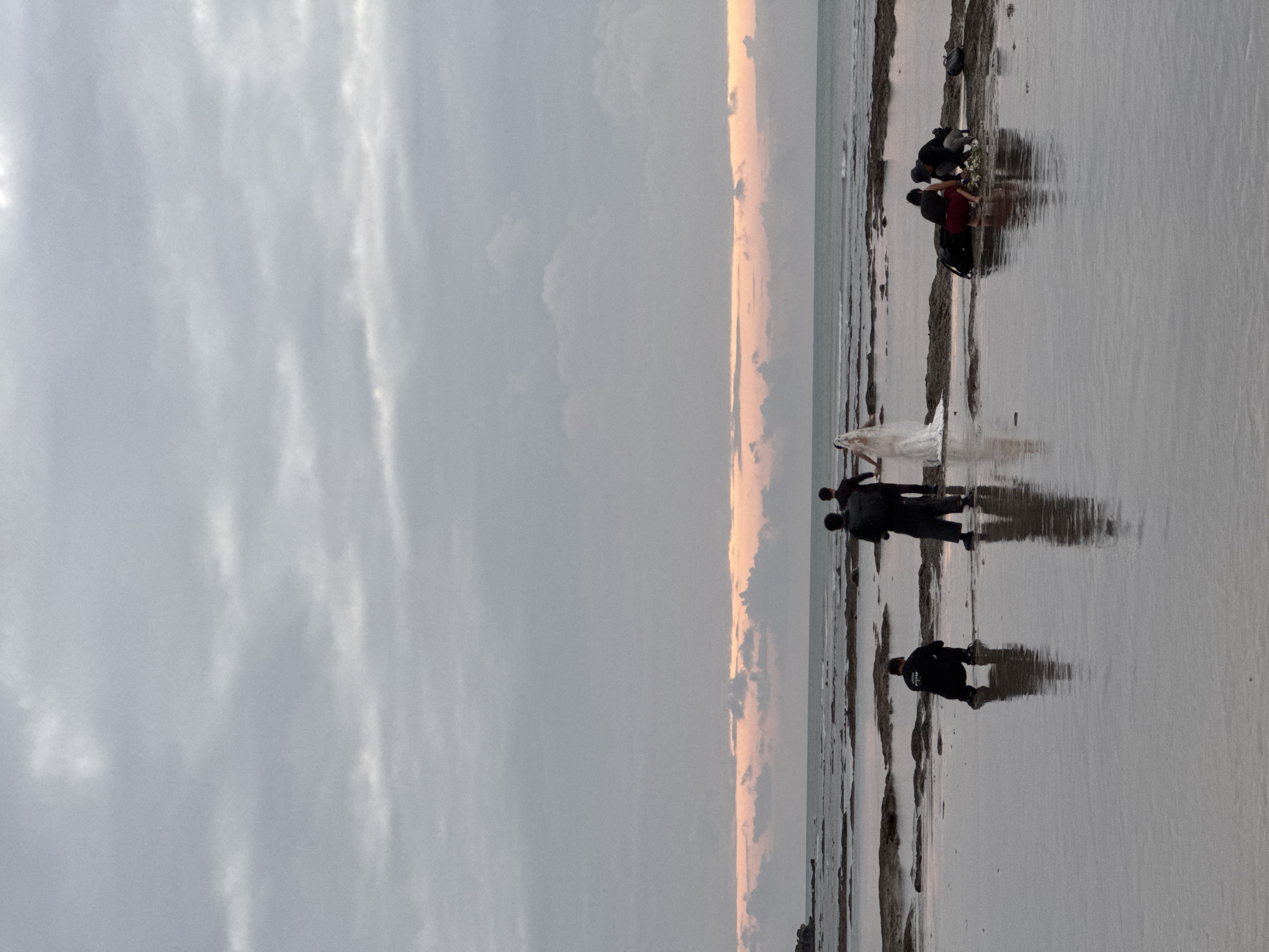 At the edge of the ocean in Kuta Selatan, Indonesia, a wedding party and their photographers are gathered on a rocky shore at low tide. The sky is overcast with hints of pink and orange from the setting sun. A bride in a white wedding dress stands with a groom and two other individuals, likely members of the wedding party. Two photographers are actively taking pictures, one crouching to get a low angle, and another standing. Two additional figures are also present, one of whom appears to be adjusting the bride's veil. The water is calm and reflects the figures and the sky.