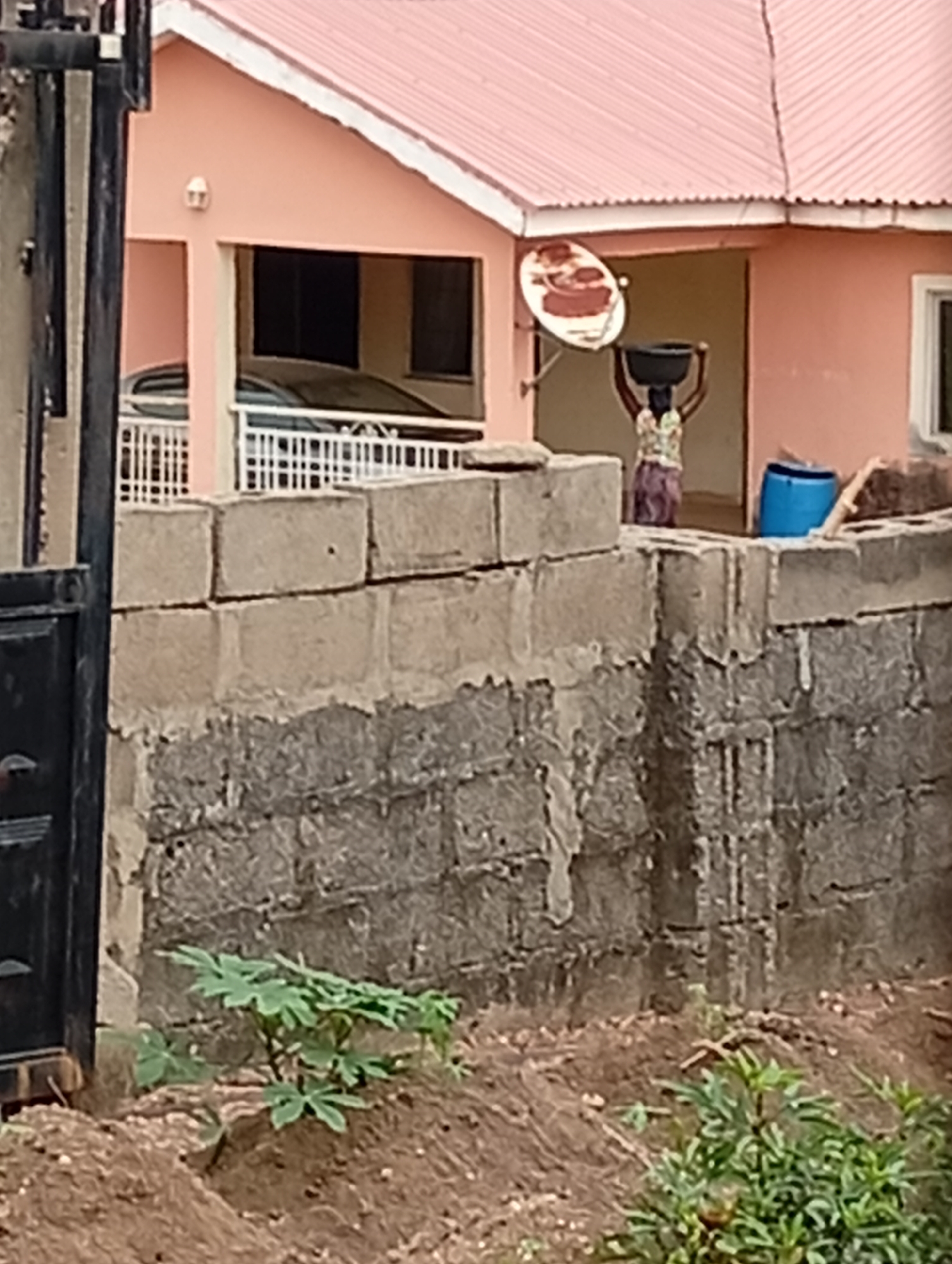 The image shows a partial view of a building in Adankolo, Nigeria, behind a concrete block wall. The building is peach-colored with a pink roof. A satellite dish is mounted on the side of the building. A woman carries a black basin on her head. A blue plastic barrel sits near her. A car is visible in the garage. The wall is made of gray concrete blocks, with a rough texture. Vegetation is present in the foreground, including a small tree and a leafy plant growing near the base of the wall. A black metal gate is partially visible in the foreground, to the left. The ground is dirt.
