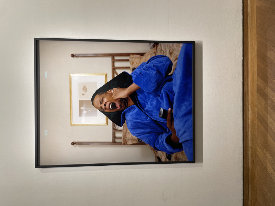 A framed photograph on a wall depicts a person in a bright blue robe yelling and holding a black object, possibly a phone, to their ear. The background suggests a bedroom setting with a four-poster bed and a framed picture hanging on the wall. The photograph is in a black frame, and the wall it hangs on is a light neutral color. To the right of the photograph is a section of patterned wooden flooring.