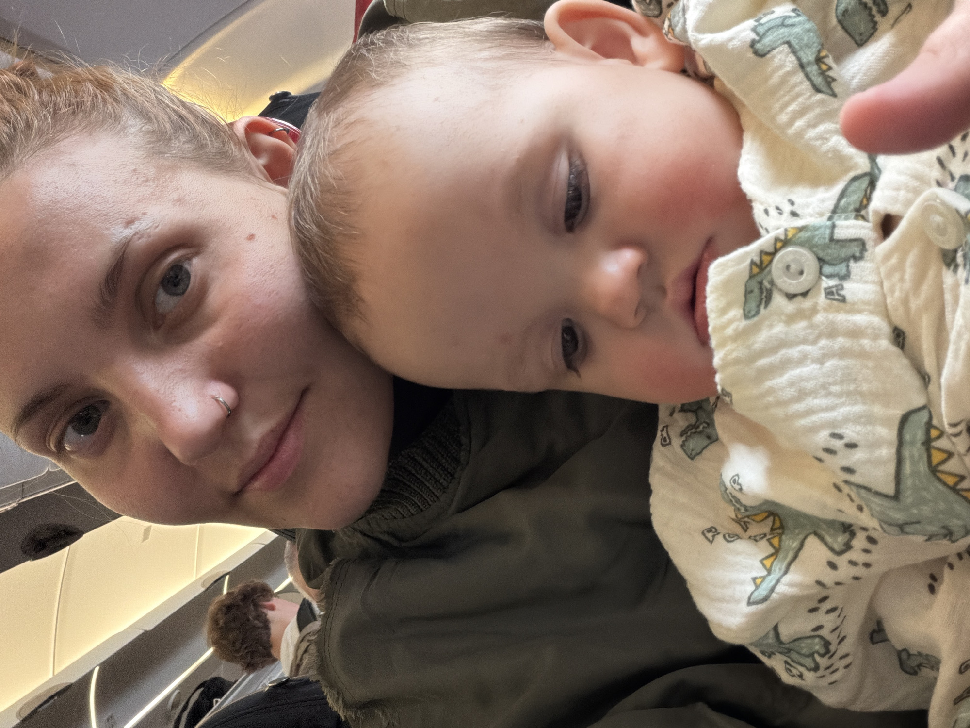 A selfie taken on an airplane shows a woman with light skin, light brown hair pulled back in a bun, and a nose ring, cradling a baby. The baby, also with light skin and light brown hair, is wearing a white outfit patterned with green and yellow dinosaurs. A portion of another passenger's head is visible in the background. The scene suggests an airplane cabin environment. There is no evidence to support the location being Khimki, Russia.