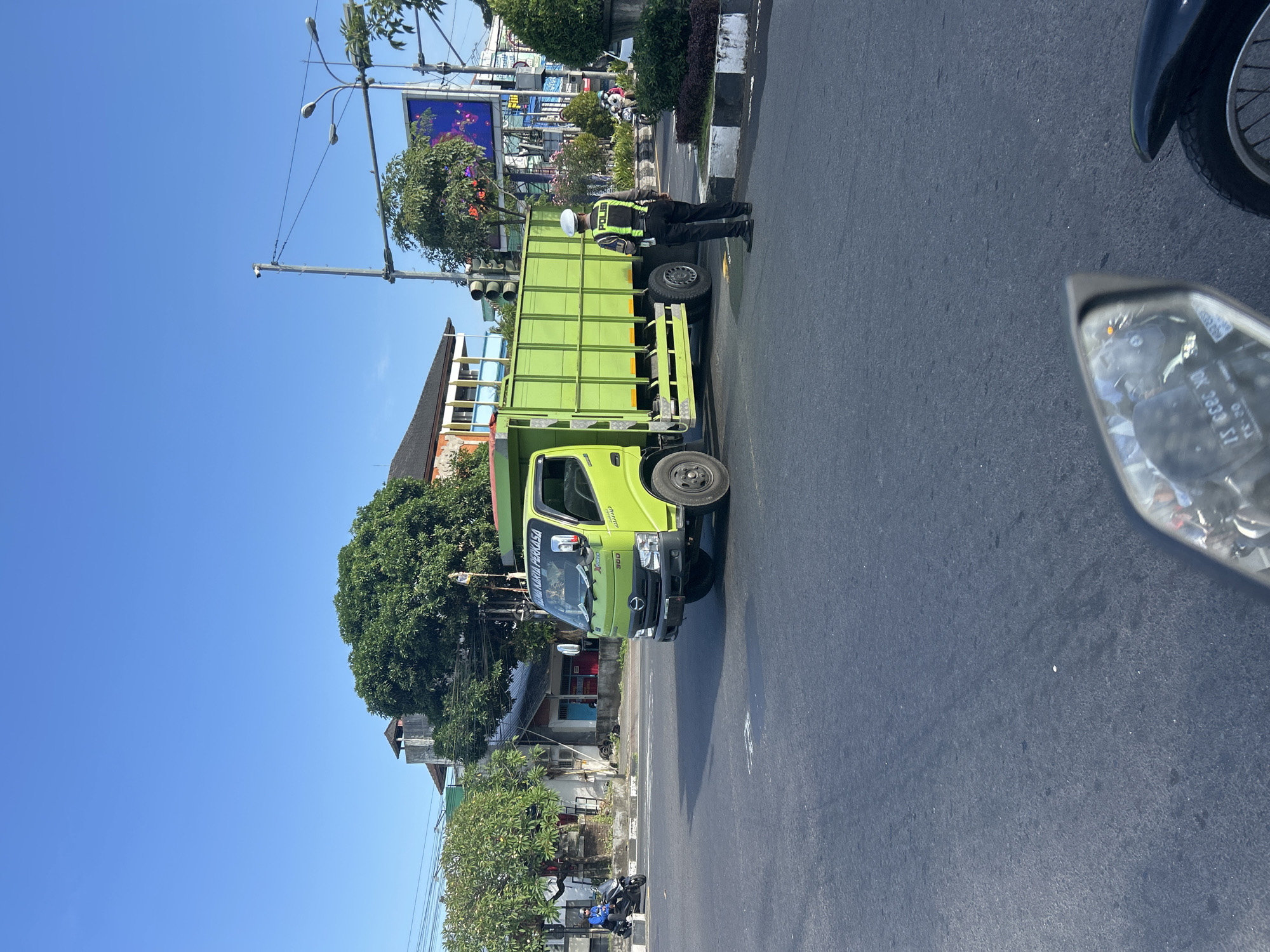 A bright green truck is stopped at a street intersection in Denpasar, Indonesia. A traffic police officer in a reflective vest and a white helmet stands to the right of the truck, facing away from the camera and observing traffic. The truck has a "HINO" logo and appears to be a medium-duty delivery truck. The intersection is paved and appears to be a typical urban street with buildings and trees lining the background. A blue sky with no clouds is visible above. The image is taken from the perspective of someone on a motorcycle, as evidenced by the reflection in the motorcycle's mirror in the foreground.