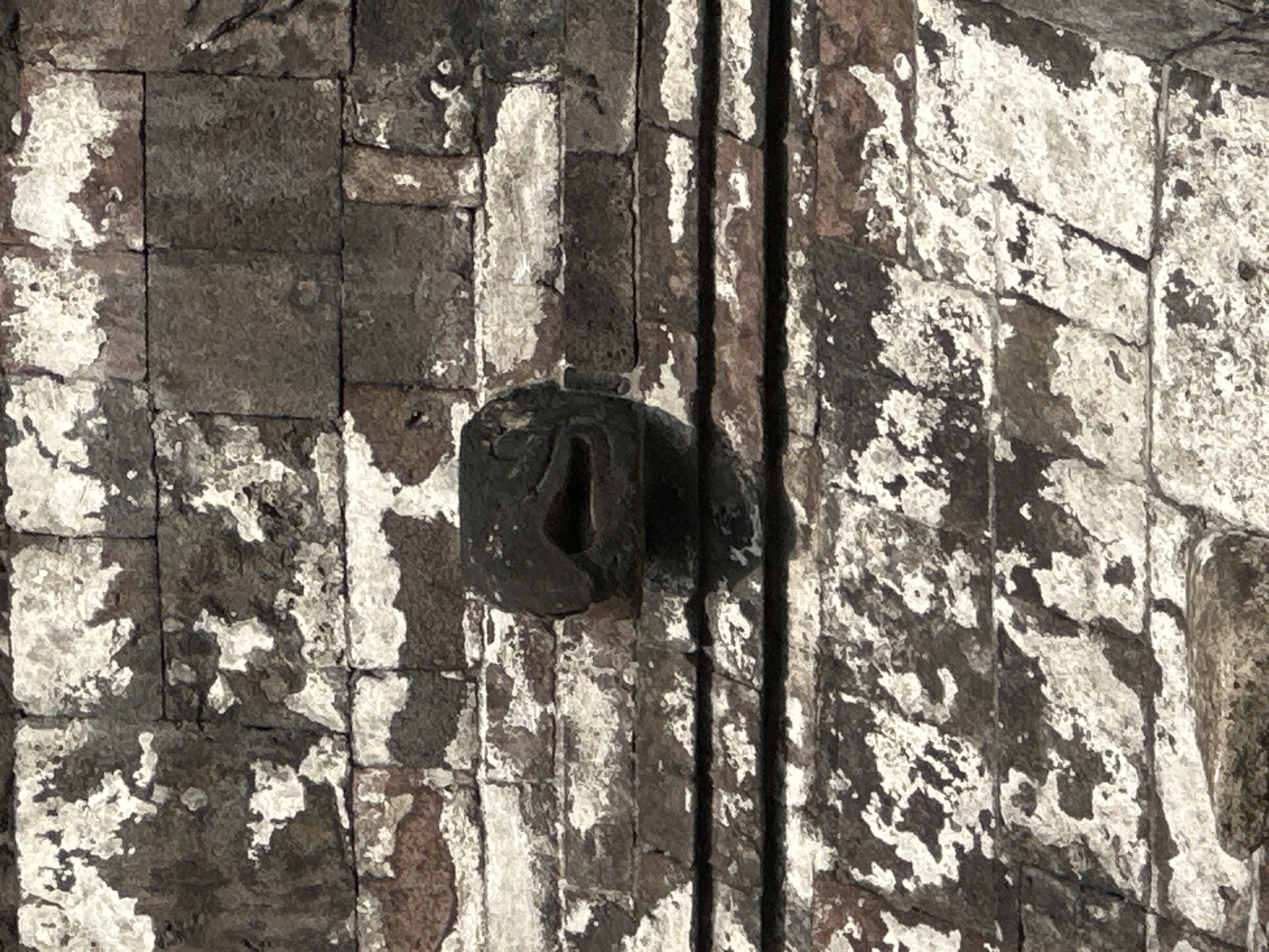 The image shows a close-up of a weathered stone wall, likely part of an ancient structure, exhibiting a mix of dark grey and lighter, faded patches. A dark, circular stone fixture with a carved or worn opening is embedded in the wall, casting a shadow. The wall is constructed with what appears to be rectangular stone blocks of varying sizes. There are vertical indentations, likely design features of the stone wall structure. The image depicts weathered stone details of ancient building in Mexico City, Mexico.