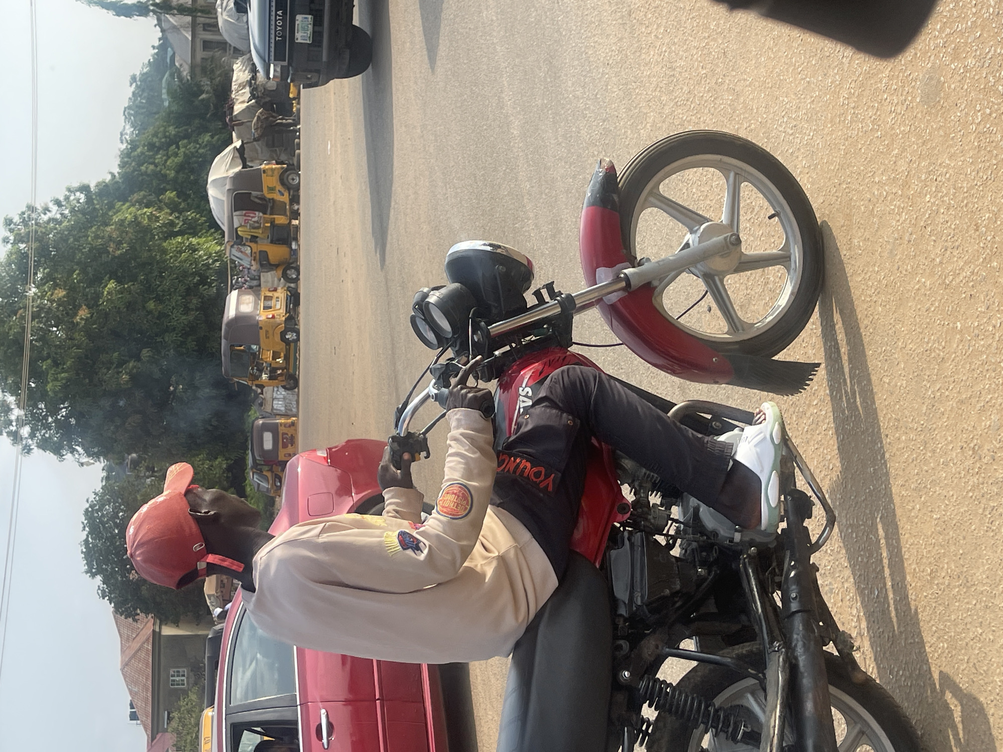 A young Black man wearing a red baseball cap, a light-colored hooded sweatshirt with patches on the sleeves, and dark pants is sitting on a red motorcycle. His right foot is resting on the motorcycle's footpeg, and he is wearing white sneakers. The man appears to be looking off to the side, away from the camera, with a neutral expression.
The motorcycle is parked next to a red car. In the background, there are several yellow auto-rickshaws, also known as "keke napep," and a black Toyota SUV. Trees and buildings are visible further in the background, suggesting an urban or semi-urban environment. The scene takes place on a paved road under a bright sky, indicating it is daytime. The lighting suggests it could be mid-morning or afternoon.
The overall impression is a candid snapshot of everyday life in a Nigerian village or town. There is no specific event or activity clearly depicted beyond the man being on his motorcycle. The presence of multiple auto-rickshaws points to a location where this is a common mode of transportation. Visible text includes "TOYOTA" on the SUV and "YOUNG" on the man's jacket. There are also some patches with text on the man's sweatshirt, but they are not clearly legible.