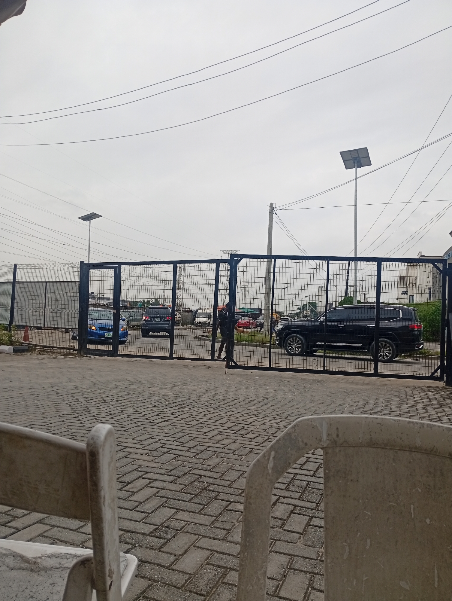 A view of a gated entrance in Ilado, Nigeria, on an overcast day. The foreground shows the backs of two weathered, light-colored plastic chairs and a paved courtyard made of interlocking grey bricks laid in a herringbone pattern. Beyond the chairs, a tall black metal gate with a grid pattern stands closed. Through the gate, several vehicles are visible, including a blue sedan on the left, a dark-colored SUV in the center, and a black SUV on the right. A person in dark clothing is seen walking near the gate in the middle distance. A chain-link fence borders the area behind the gate, and in the far distance, power lines and utility poles crisscross the sky above buildings and vegetation.