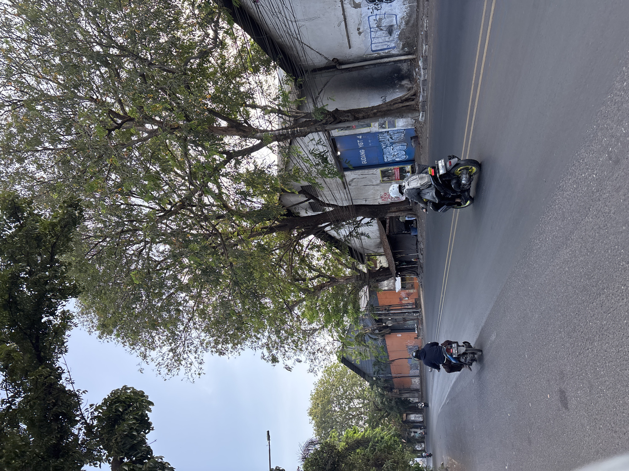 The image depicts a street scene in Cirebon, Indonesia, characterized by mature trees lining the roadway and industrial-style buildings with weathered facades. Two individuals are visible riding motorcycles on a paved road. One motorcycle, in the foreground and slightly to the right, has a rider wearing a white helmet and a backpack, with yellow accents on the wheels of the motorcycle. Another motorcycle, further in the distance and to the left, has a rider wearing a dark hooded jacket and a dark-colored sarong or lower garment.
The buildings alongside the road appear to be warehouses or industrial structures, some with graffiti and weathered paint. One prominent building features a large blue door with text that reads "GUDANG YGT 4". Power lines are strung across the top of some of the buildings. The sky is clear and bright, suggesting daytime. The overall atmosphere is that of a functional urban environment with a mix of natural elements (trees) and man-made structures.