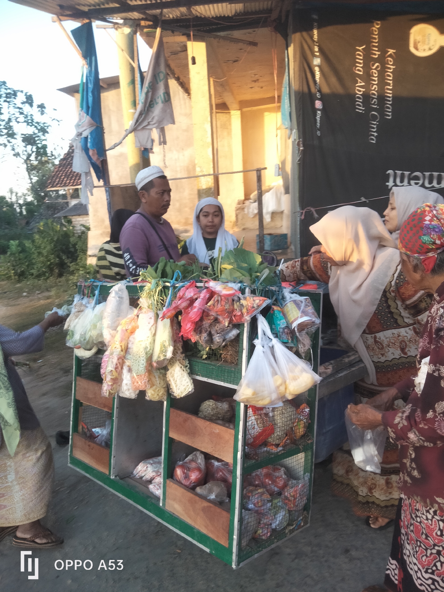 This image depicts an outdoor market scene in the city of Badung, Indonesia, likely in the late afternoon given the warm lighting. At the forefront, a mobile market stall displays various goods, including packaged snacks and fresh produce like leafy vegetables and what appear to be tomatoes. Several individuals are present around the stall. A man wearing a white kufi and a purple shirt is standing behind the stall, seemingly engaged in transactions. Next to him, a young woman in a white hijab is also behind the stall. To the right, a woman in a light-colored hijab and an ornately patterned dress is interacting with the stall, holding a plastic bag. Further to the right, another elderly woman, adorned in a dark red patterned top and a batik-patterned skirt, is also near the stall. A person on the far left, wearing a patterned headscarf and a skirt, has their arm extended towards the stall, possibly making a purchase. The stall itself is constructed of wood and metal mesh, with multiple shelves holding the merchandise. In the background, a building with a corrugated metal roof and textured walls is visible, along with vegetation and trees. A large black banner with white text is hanging behind the stall, adding to the market ambiance. The ground surface appears to be paved or dirt. A watermark "OPPO A53" is present in the bottom left corner of the image.