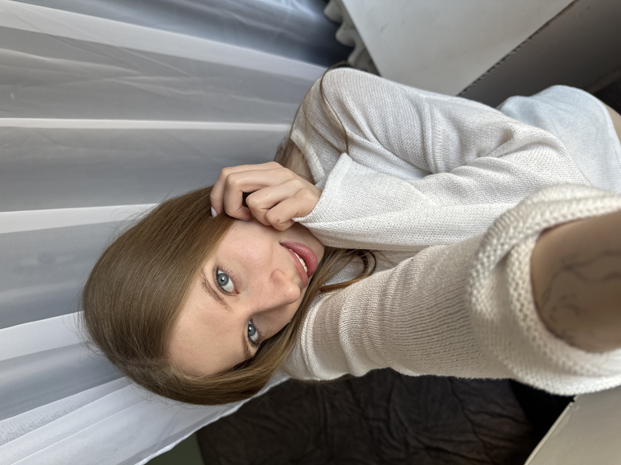 A woman with light brown hair and blue eyes is taking a selfie. She is wearing a white sweater. The background includes white blinds and a dark brown surface. No specific details indicate the location is Chaikovsky, Russia.