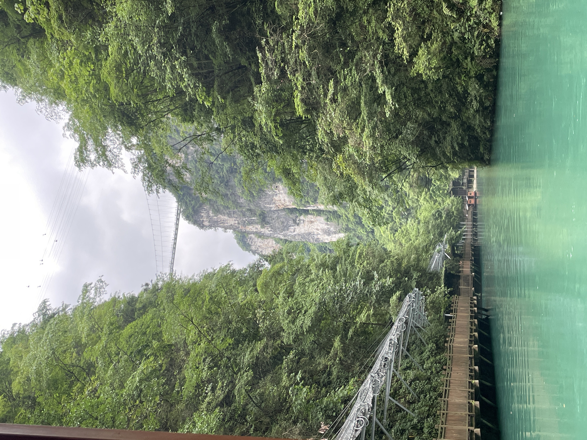 The image shows a scenic view in Zhangjiajie, China, featuring a turquoise river bordered by lush green vegetation. A wooden walkway extends along the water's edge, with metal structures supporting a higher path or cable system. In the background, a suspension bridge stretches across a valley, connecting to a rocky cliff face partially obscured by clouds. The environment suggests a mountainous and forested area. There are no visible people in the immediate scene.