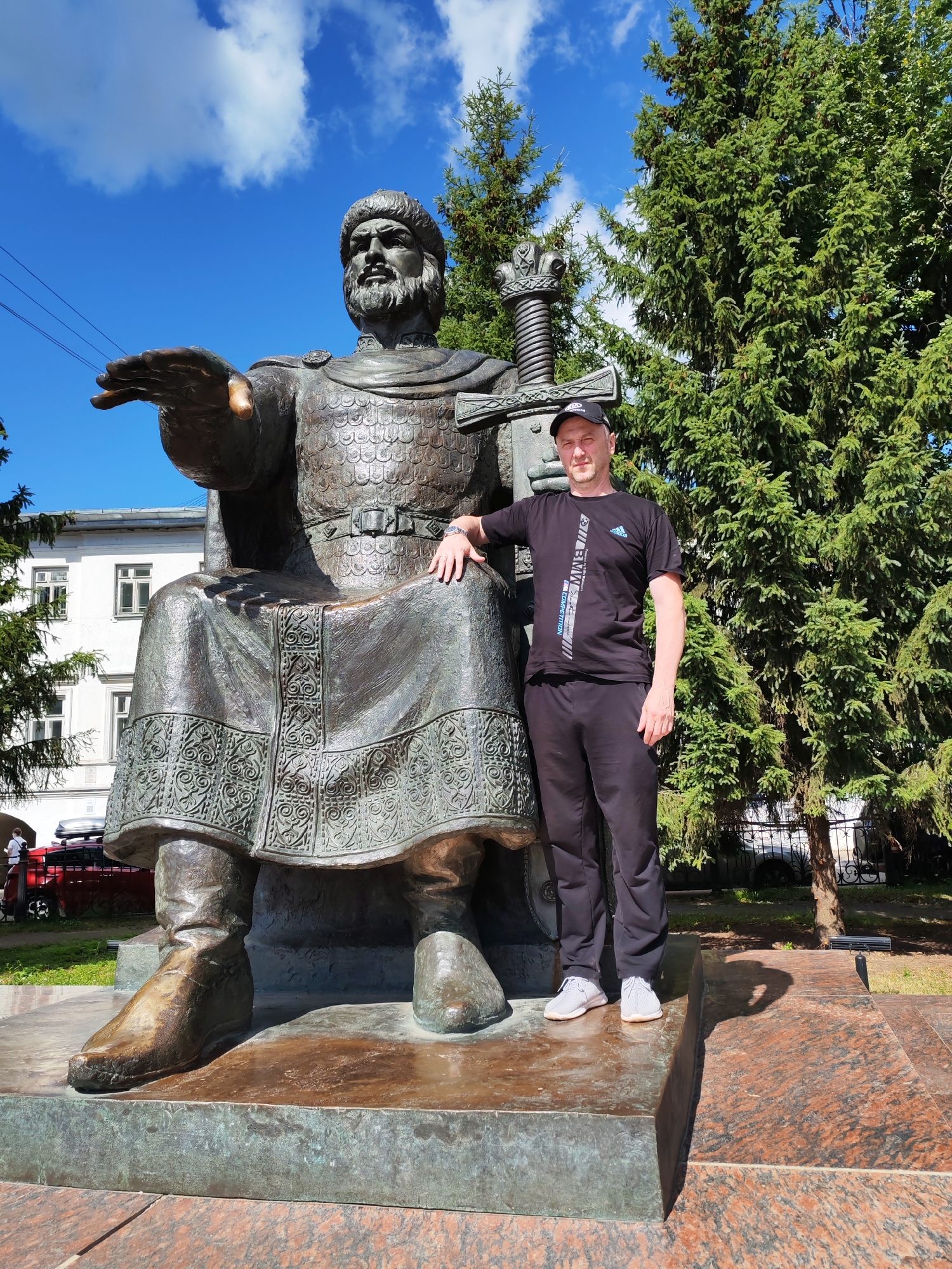 This media file shows a man standing next to a large bronze statue of a seated historical figure. The man is wearing a black t-shirt with white and blue lettering, black pants, a black baseball cap, and white sneakers. He is smiling and has his right hand resting on the statue's knee and his left hand on the statue's arm. The statue depicts a man with a beard, wearing armor and a fur-trimmed hat, with a large sword resting behind his shoulder. The statue is seated on a large stone base, with intricate carvings on its skirt and boots.
The scene is set outdoors on a sunny day, with a clear blue sky and some white clouds visible. Tall evergreen trees are in the background, and a white building with windows is also visible behind the statue. The ground around the statue is paved with red and grey granite tiles. The overall impression is a bright, clear day, likely during daylight hours, in a public space such as a park or square. The man appears happy and proud to be posing with the statue. The location is identified as Kostroma, Russia. There is visible text on the man's t-shirt.