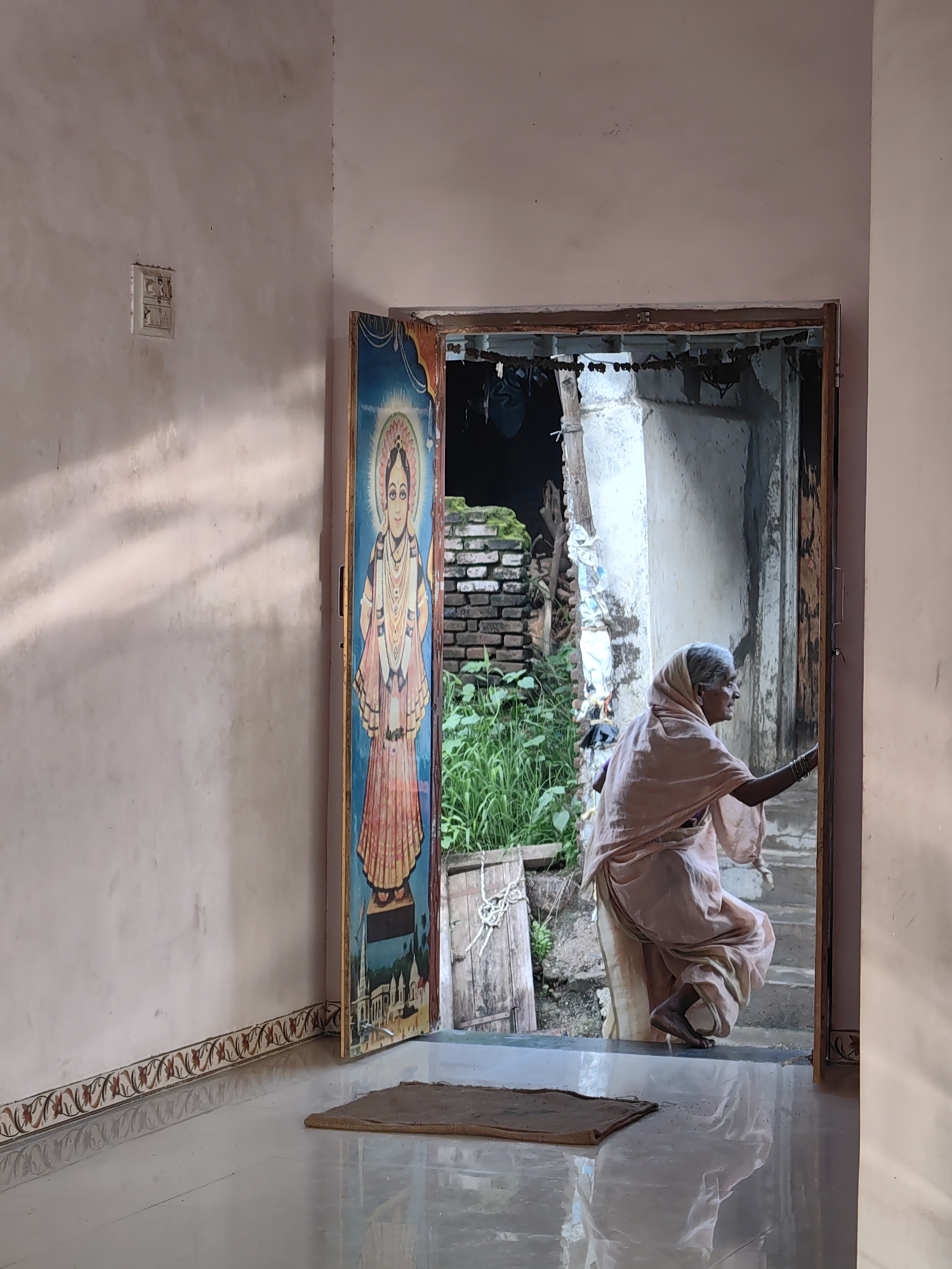An elderly Indian woman, wearing a light pink sari and head covering, is captured in a doorway in Mhaiswadi, India. She is positioned mid-stride, her body angled towards the right as she appears to be stepping through the doorway. Her right hand is visible reaching towards the doorframe. The doorway itself is framed by a weathered wooden structure, with what appears to be a painted religious mural on the inside of the left door panel, depicting a figure in traditional attire. Beyond the doorway, an outdoor scene is visible, including rough brickwork, lush green foliage, and a rustic wooden structure. The floor inside the doorway is tiled and reflects the light. To the left of the doorway, on a light pink wall, a light switch is mounted. The overall setting suggests a residential entrance in a rural or semi-urban Indian environment.