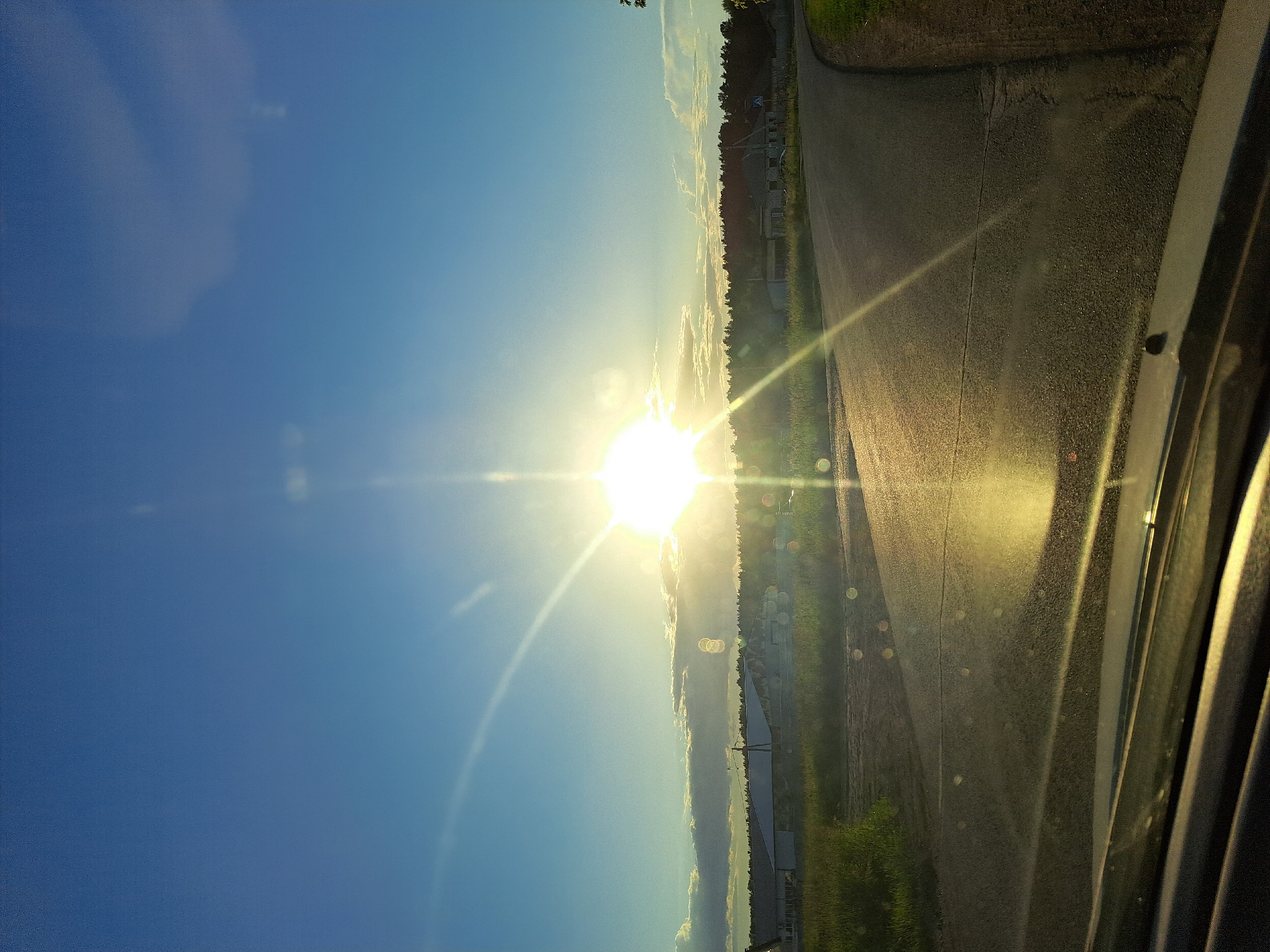 This image captures a road winding through a rural landscape under a bright, sunlit sky. The sun is positioned high and to the left of center, emitting strong rays and creating lens flare effects on the camera. The road is paved and appears to be in good condition, with a light-colored line marking its center. To the left of the road, there is a grassy field with some sparse vegetation. Beyond the field, a line of trees forms a dark silhouette against the horizon. In the distance, there are several buildings, including what appear to be farm structures or warehouses, one with a distinct white roof. The sky is a clear, vibrant blue with a few wispy clouds on the horizon, catching the sunlight. The foreground of the image shows the hood and windshield of a vehicle, suggesting the photo was taken from inside a car. The overall scene depicts a sunny day in a rural setting.