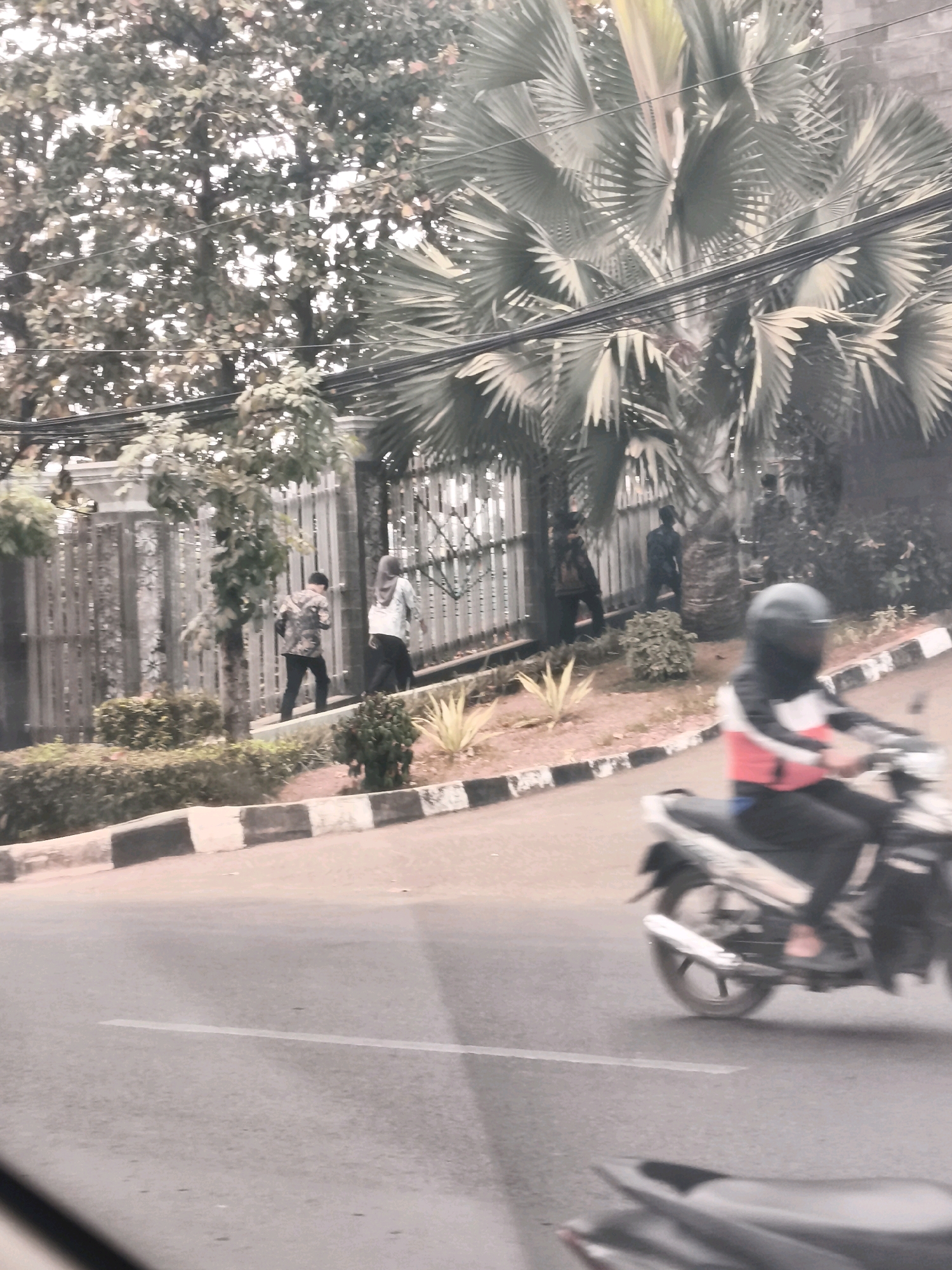 The image shows a street scene in Palembang, Indonesia. In the foreground, a person on a motorcycle is riding on the right side of the road. The rider is wearing a helmet, a red and white jacket, and dark pants. The motorcycle is silver and black. In the background, several people are walking along a sidewalk next to a decorative metal fence. One person is wearing a camouflage jacket and dark pants, another is wearing a light-colored hijab and dark pants, and a third is wearing dark clothing. Behind the people, there is lush vegetation, including palm trees and smaller bushes. The road has a white line marking the lane. The overall scene appears to be a typical day in a city with moderate traffic and pedestrian activity.