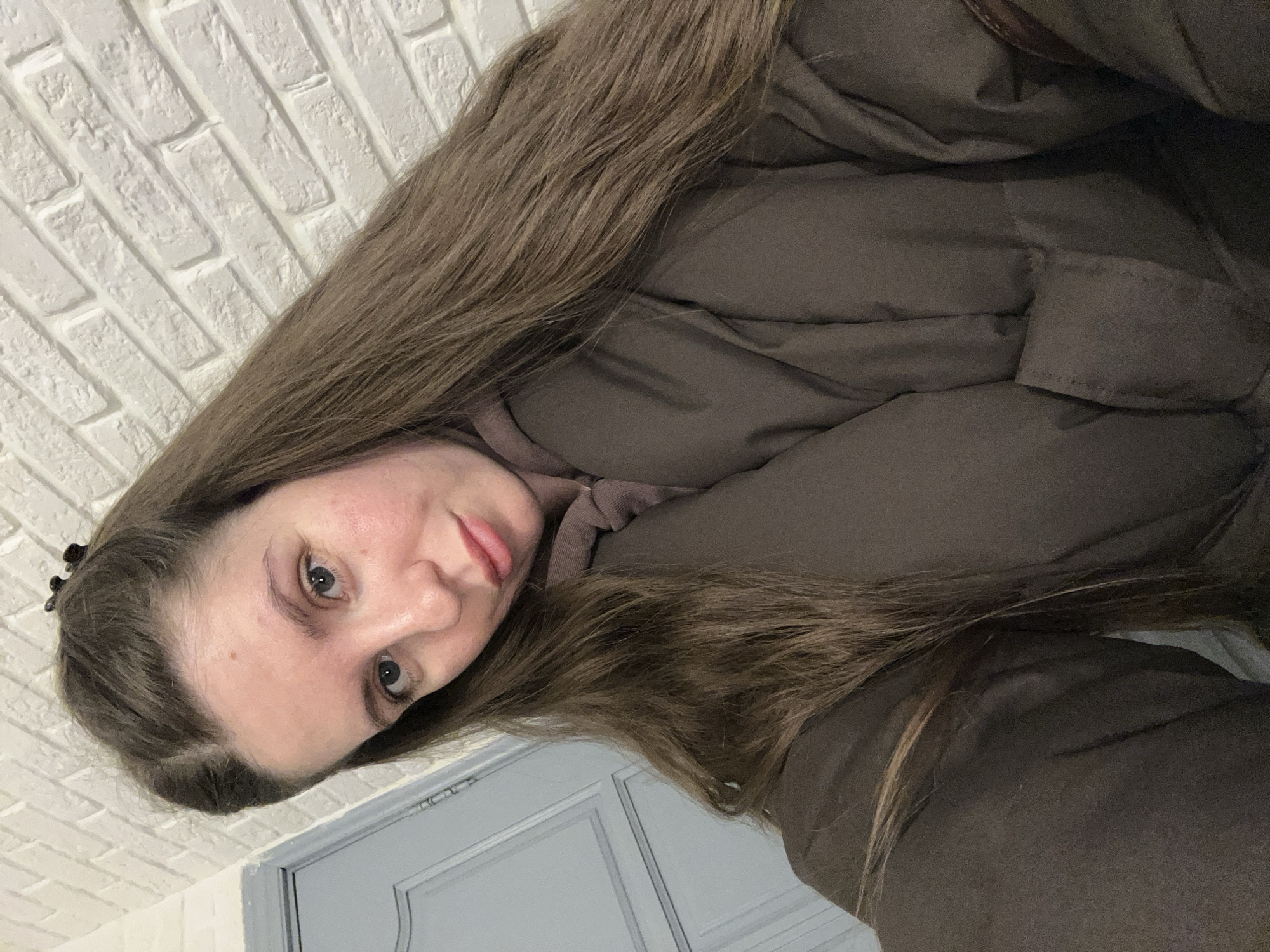 A woman is in an indoor setting with a white brick wall visible behind her on the left and a gray door partially visible at the bottom of the frame. She is wearing a brown, puffy winter coat with a hood and has long, brown hair. The location is Klin, Russia.