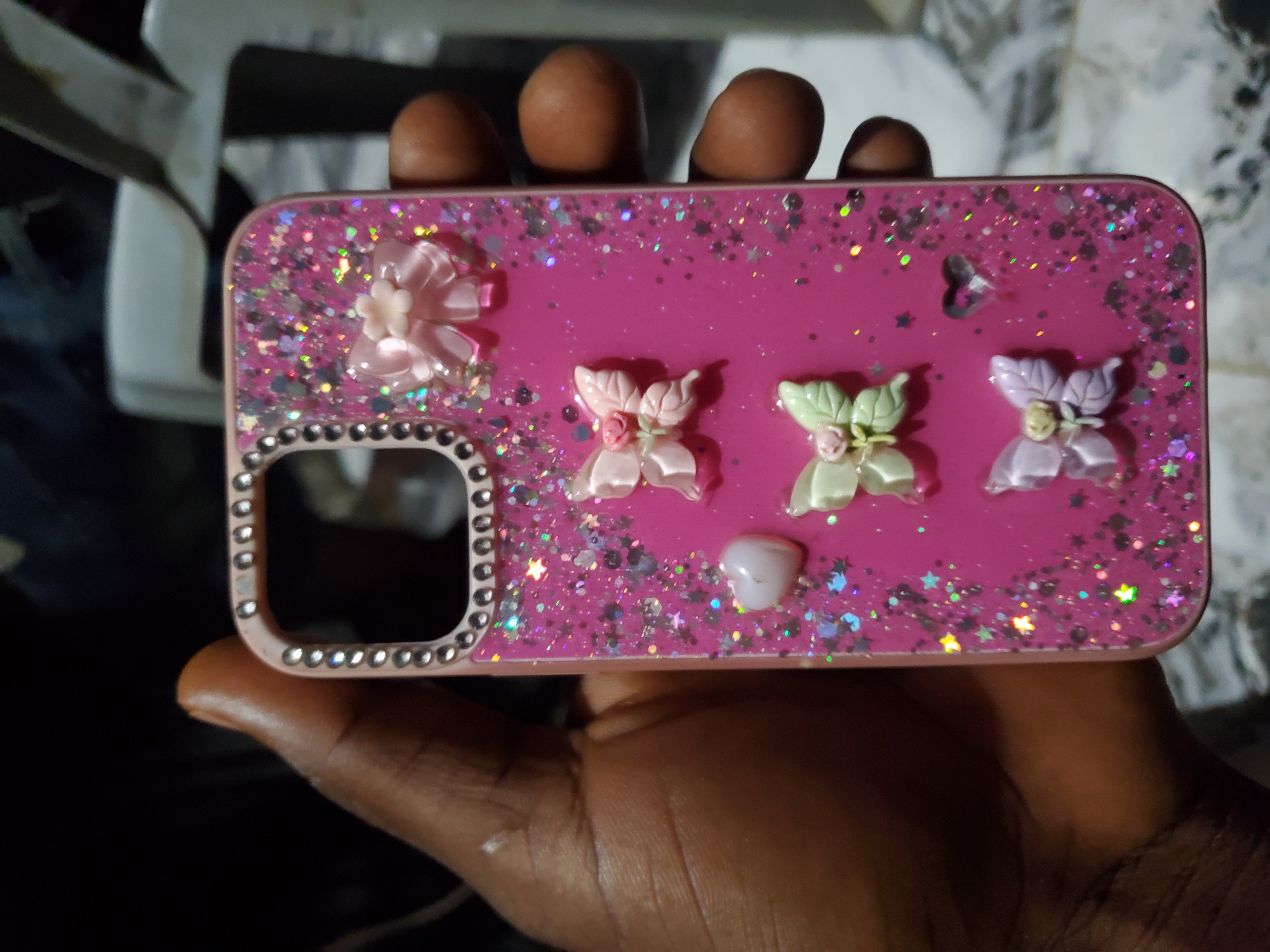 A dark-skinned hand holds a hot pink mobile phone case, positioned vertically, with the bottom facing down and the camera cut-out at the top-left. The case features a clear, liquid glitter-filled back, containing silver and iridescent star-shaped and circular glitter pieces. The camera cut-out is bordered with small, clear rhinestones. Decorative appliqués are adhered to the case's surface: a light pink bow with a white center flower is at the top-right; below it, a light pink butterfly with a pink rose center; further down, a light green butterfly with a pink rose center; and at the bottom-right, a light purple butterfly with a cream rose center. A small, white, heart-shaped appliqué is situated mid-left, and a clear heart-shaped element is visible near the bottom-right. The background is out of focus, indicating an indoor setting with indistinct objects and varying light levels.