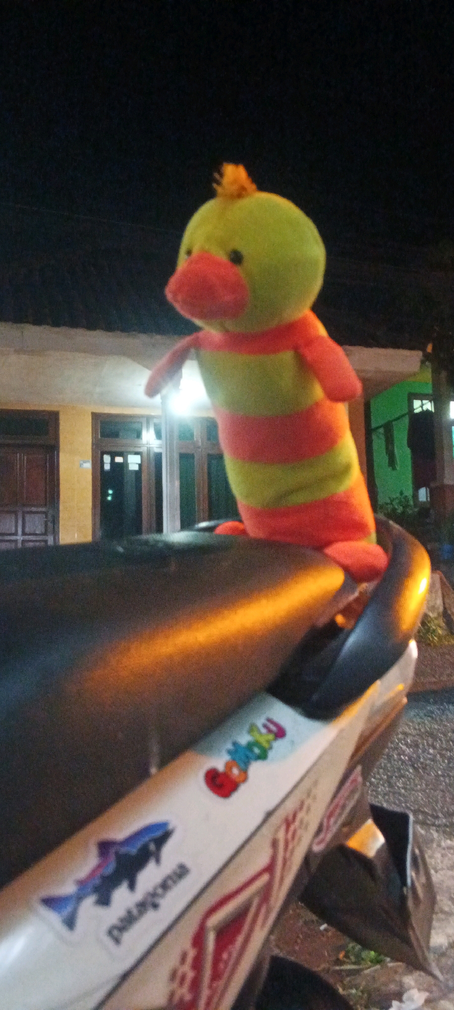 In the city of Kesiman, Indonesia, at night, a plush toy of a green duck-like creature with orange stripes and a red beak stands upright on the seat of a dark-colored motorcycle. The background shows the facade of a building with yellow walls and brown doors, illuminated by lights from within the building and possibly from outside streetlights. A portion of a green wall is visible to the right of the building. The motorcycle seat has two stickers: one is a blue silhouette of a leaping fish with the word "patagonia" below it, and another sticker with colorful letters that appears to say "Gotoku." The image is slightly blurry due to low light conditions and possibly motion.