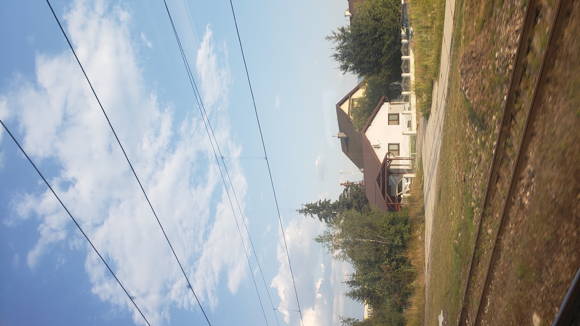 The image depicts a view from a moving train, as indicated by the blurred foreground of railroad tracks and the presence of overhead wires. The scene is set in Liptovská Teplá, Slovakia, under a partly cloudy sky. In the mid-ground, several residential houses are visible, nestled amongst trees and vegetation. One house in particular is a white structure with a brown roof and a covered carport sheltering a white car. Adjacent to it is another house with a yellow facade and a darker brown roof. A white fence encloses a portion of the property. The surrounding area features a mix of greenery, including trees and grassy areas, with a pile of dirt or debris in the foreground. In the background, a tall industrial structure with a red top is visible, suggesting proximity to an industrial zone or a local landmark. The overall impression is a glimpse of a suburban or semi-rural landscape.