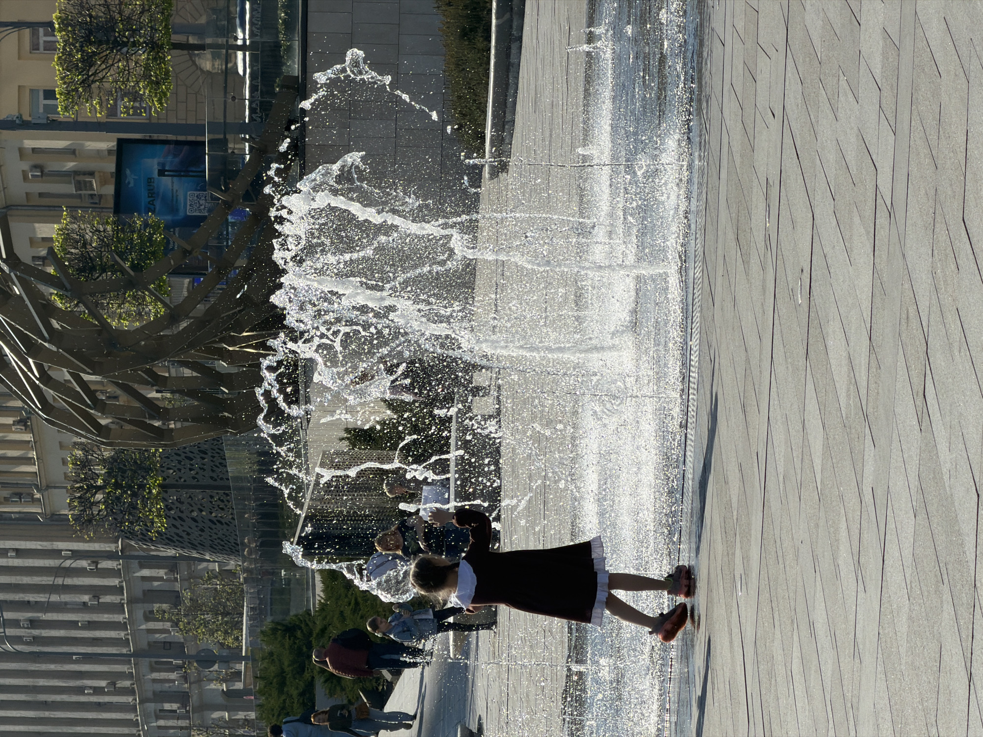 A horizontal shot depicts a sunlit urban scene. A girl in a dark red dress with a white collar and cuffs, wearing reddish shoes, stands facing the water, seemingly interacting with it in the foreground. A modern fountain releases water in various directions, creating a dynamic, splashy effect. Other people are visible in the background, some appearing to be watching or interacting with the fountain as well. To the left of the fountain, there is a metal sculptural structure, behind which buildings are visible. There is also green trees present behind the metal. The ground is paved with light grey stones.
This appears to be the Zaryadye Park and the "Soaring Bridge" fountain in Moscow, Russia.