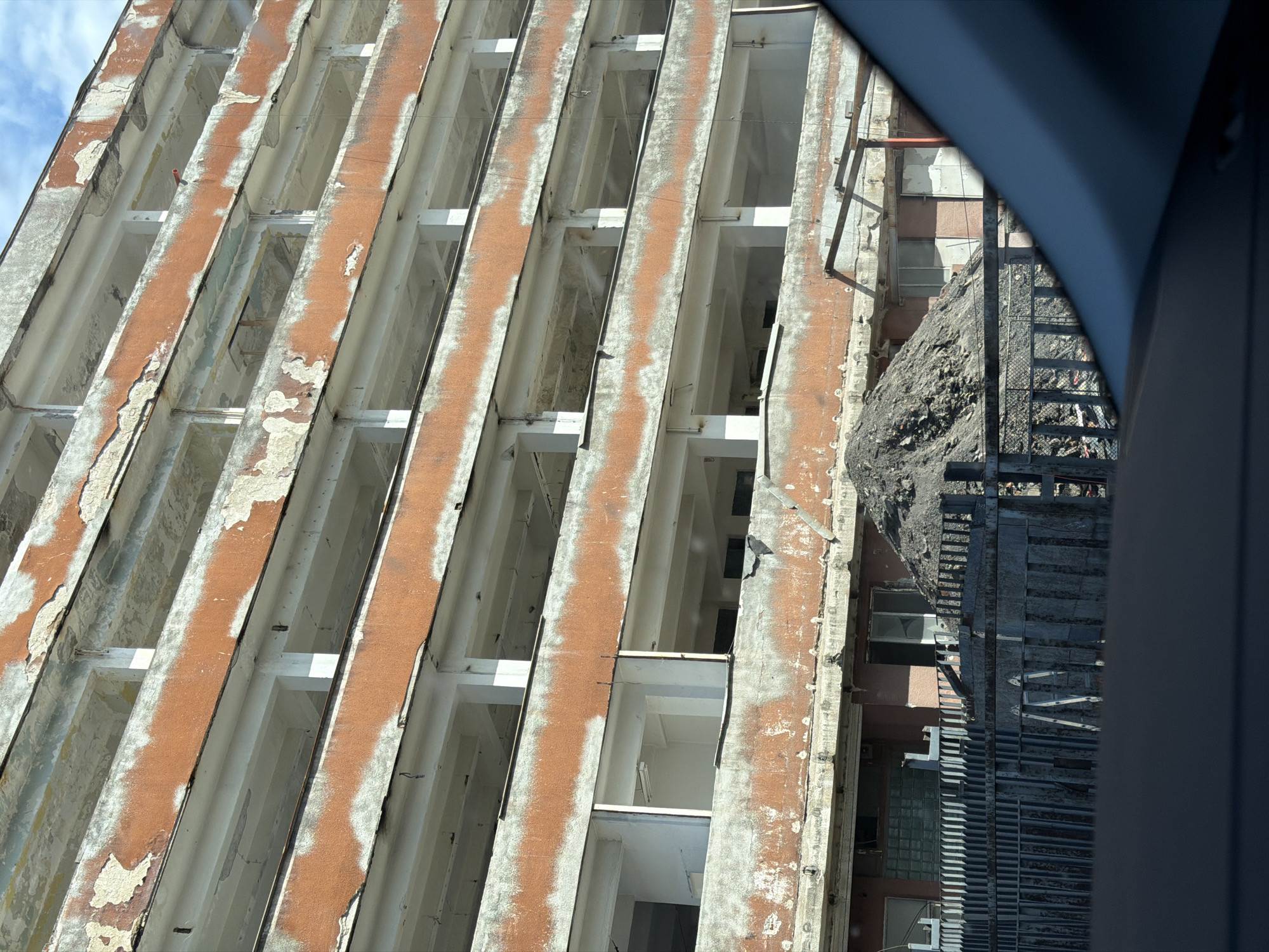 The image shows a partial view of an unfinished or dilapidated building in Plovdiv, Bulgaria. The facade consists of multiple floors with repetitive rectangular openings where windows would typically be. The building's structure features alternating horizontal bands of orange and white, with the orange bands showing signs of peeling paint and deterioration. An external metal fire escape runs alongside the building, partially obscured by a debris pile. The view is framed by an out-of-focus dark shape in the foreground, possibly from a window or other structure. The sky is partly visible in the upper left corner, suggesting a daytime setting.