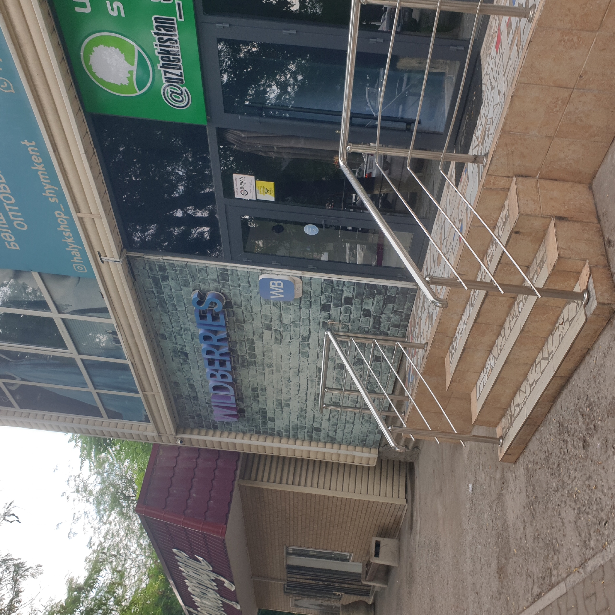 The image shows the exterior of a Wildberries store, a Russian online retailer, located in the city of Shymkent, Kazakhstan. The storefront features a modern design with a brick-patterned facade and large glass doors. Above the entrance, the "Wildberries" logo is prominently displayed. To the right of the entrance, a green sign with a white tree logo and the text "@uzbekistan" is visible. The store is accessed via a set of tiled stairs with metal railings. In the background, trees and other buildings can be seen. There are no people or ongoing actions depicted in the image.