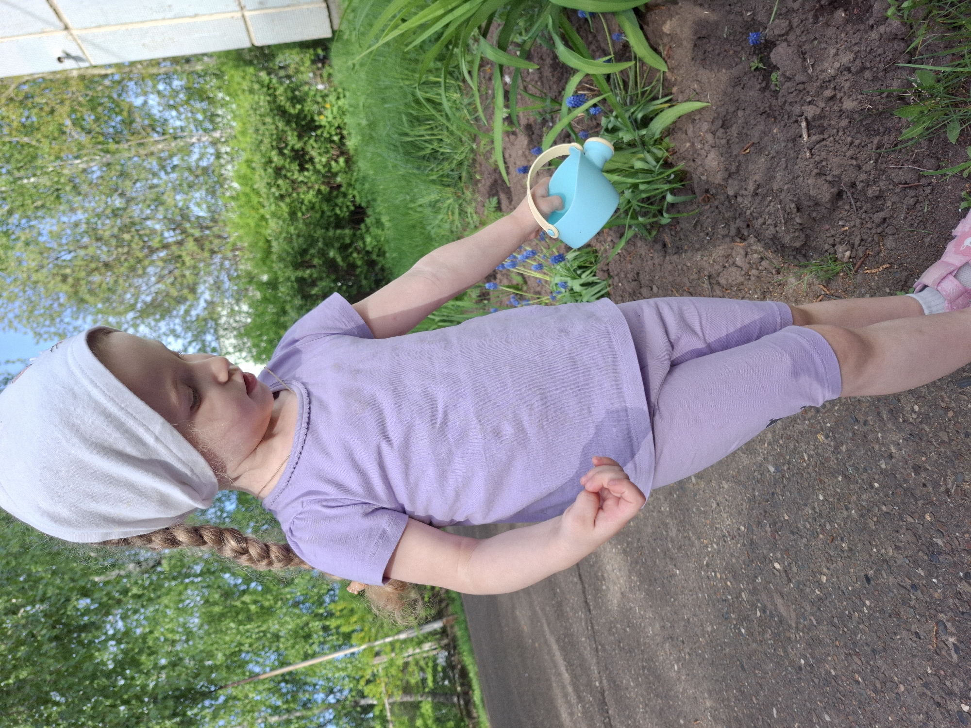A young child, likely female, is seen outdoors in Chaikovsky, Russia, near a garden. The child is wearing a light purple t-shirt and matching shorts, a white head covering, and pink sandals with white socks. Her hair is braided. She holds a small, blue watering can in her right hand, positioned over a flower bed containing green plants and small blue flowers. The ground is dark soil. The child appears to be watering the flowers. The background includes a concrete path, green foliage, and parts of a building.
