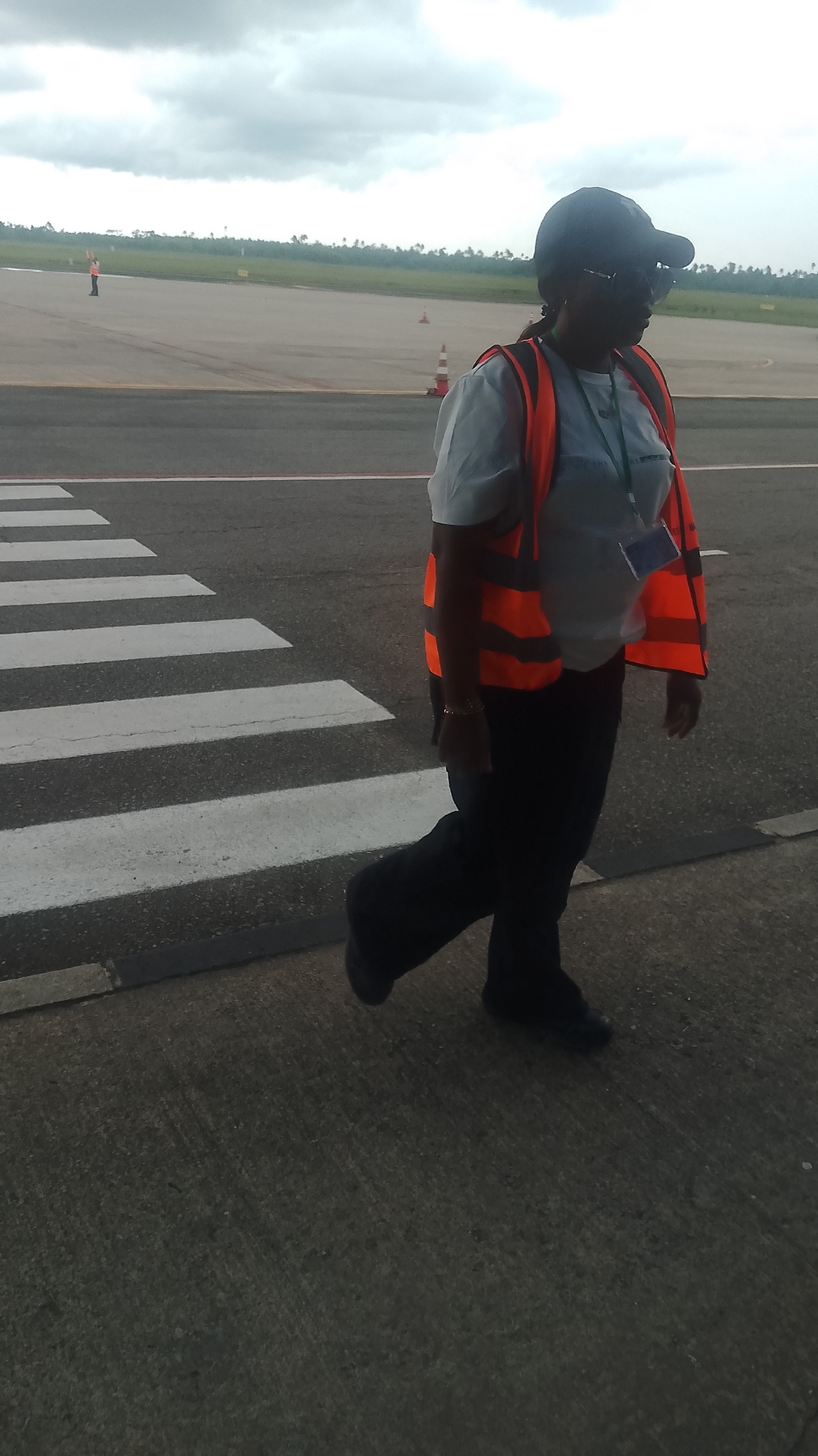 A woman wearing a dark baseball cap, sunglasses, and an orange high-visibility vest over a light blue shirt walks across a pedestrian crossing at an airport. She is also wearing dark pants and dark shoes. The pedestrian crossing has thick white stripes. In the background, an empty runway is visible, along with orange traffic cones, and distant greenery and trees under a cloudy sky. The woman is the only person clearly depicted in the foreground. The scene appears to be at the airport in Orerokpe, Nigeria, given the context.