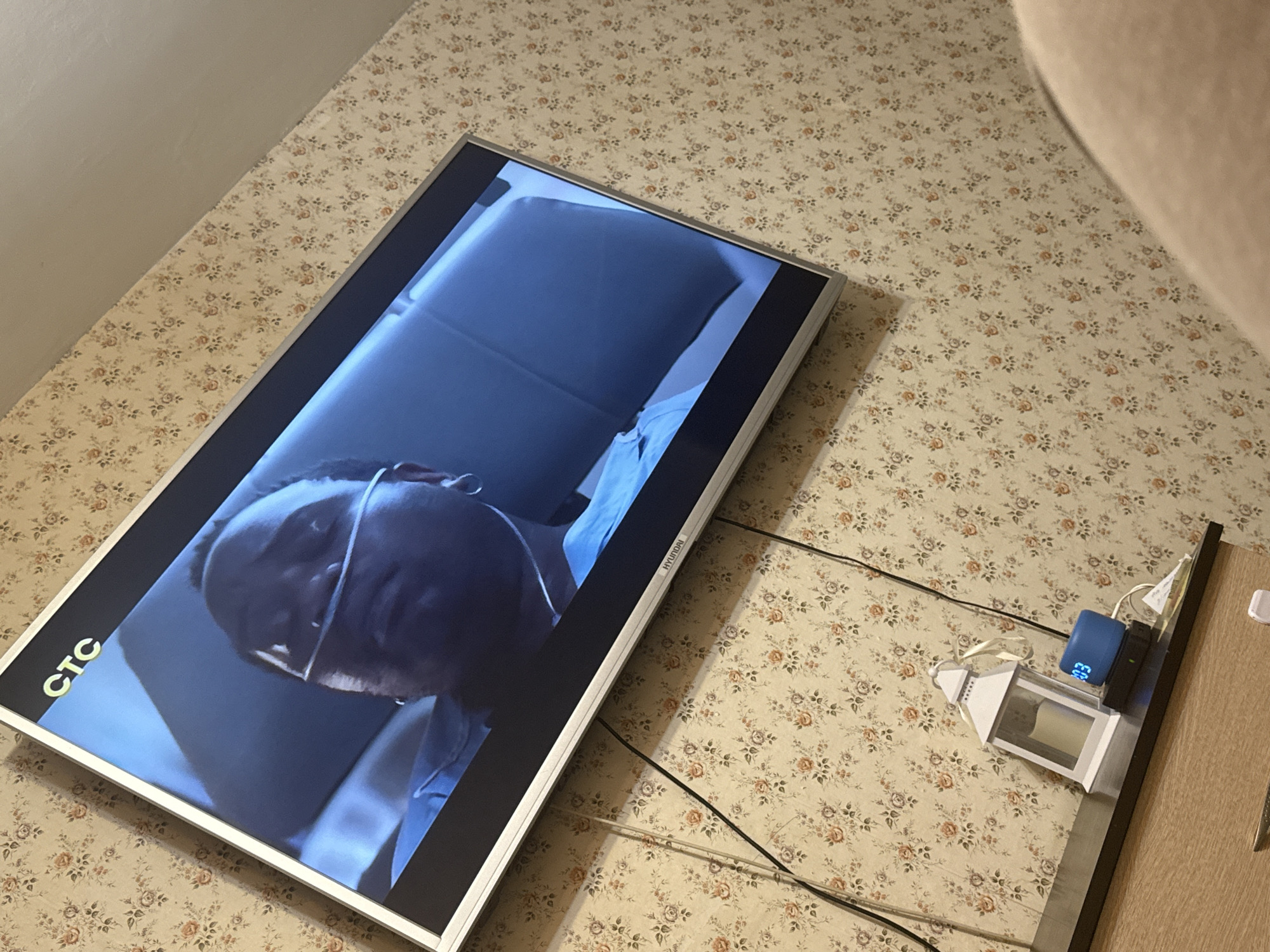 A television is showing a person wearing glasses in a dark environment. The TV is resting on a surface with a floral pattern, and several electrical cords are connected to it. Next to the TV, there is a table with a lantern and a small blue device displaying "203". The location is implied to be Mytishchi, Russia, based on the request, but there are no visible elements in the image to confirm it.