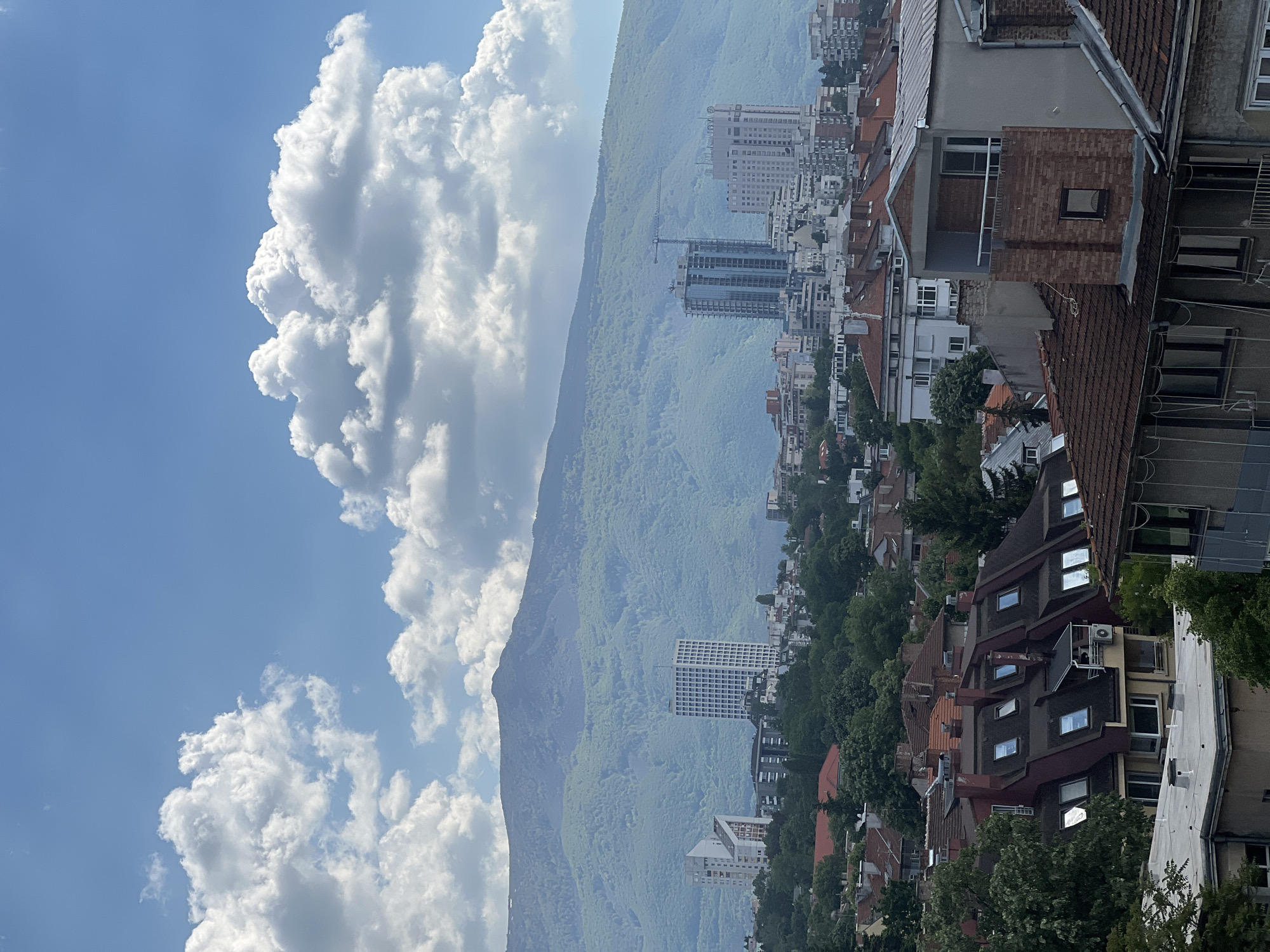 The image shows an urban landscape in Sofia, Bulgaria, likely a residential area viewed from an elevated position. In the foreground, there are dense groupings of residential buildings, mostly with reddish-brown tiled roofs, interspersed with trees. Some of the buildings have more modern features, with windows and balconies visible. Behind these structures, there is a large mountain range covered in trees, extending across the frame. The mountainside is bathed in a somewhat hazy light, giving a soft, slightly blurred appearance. In the background, there are high-rise buildings scattered among the trees and lower buildings, suggesting a mixed urban and natural environment. A construction crane is visible. The sky, covering much of the left side of the image, is blue with large, puffy white clouds. The overall orientation of the picture is vertical, and the scene appears to be captured in daylight.