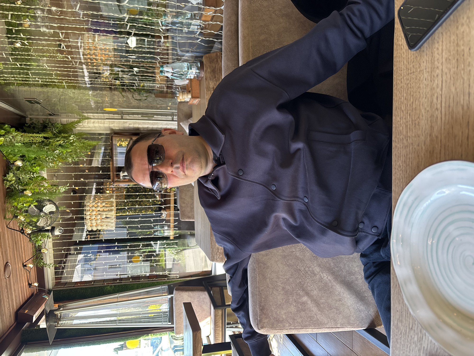 A man is seated at a wooden table in what appears to be a cafe or restaurant setting in Sochi, Russia. He is wearing sunglasses and a dark-colored, long-sleeved jacket with snap buttons, paired with dark pants. He looks directly towards the camera with a neutral expression.
In front of him on the table, a light-colored plate with a light blue or green spiral pattern is partially visible on the right. A dark-colored smartphone with a textured case rests near his right hand. The man is seated on an upholstered chair with a light brown or tan finish.
The background features large glass windows or partitions, through which abundant green plants and foliage are visible, including ferns and bushes. String lights are strung horizontally across the glass, creating a festive ambiance. Reflections of the interior, including another table and chairs, are visible on the glass.
Further into the background, a black fan and a tall, silver outdoor heater are present, suggesting the space might be a semi-outdoor or adaptable area. A decorative light fixture resembling a basket filled with spherical lights hangs from above. Another table with a clear bottle of liquid can be seen behind the man. The lighting suggests it is daytime, possibly late afternoon, with ambient light. No discernible text is visible in the image.