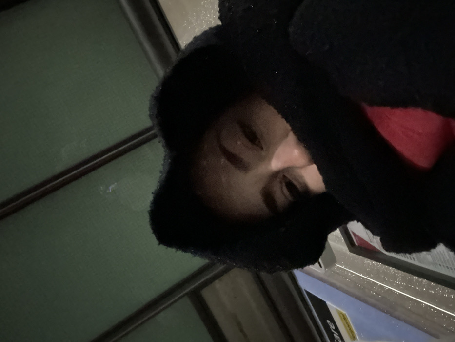 An individual is depicted in a low-angle, close-up photograph, presumed to be a selfie, taken at a public transport stop in Hœnheim, France. The subject, with medium-dark skin tone, presents their upper face, including eyes, nose bridge, and forehead. The lower portion of the face is obscured by a black, fluffy garment, which appears to be a hood with ear-like protrusions. A segment of red clothing is visible beneath the black outerwear. The background features the underside of a green public transport shelter roof with dark horizontal structural beams. To the left, a white and yellow panel of a public transport ticket machine or validator is partially visible, displaying the text "are," likely part of "Fare" or "Tarif." The ambient light indicates a low-light environment, possibly nighttime, with speckles of light on background surfaces suggesting reflections or moisture. The individual's eyes are directed towards the camera.