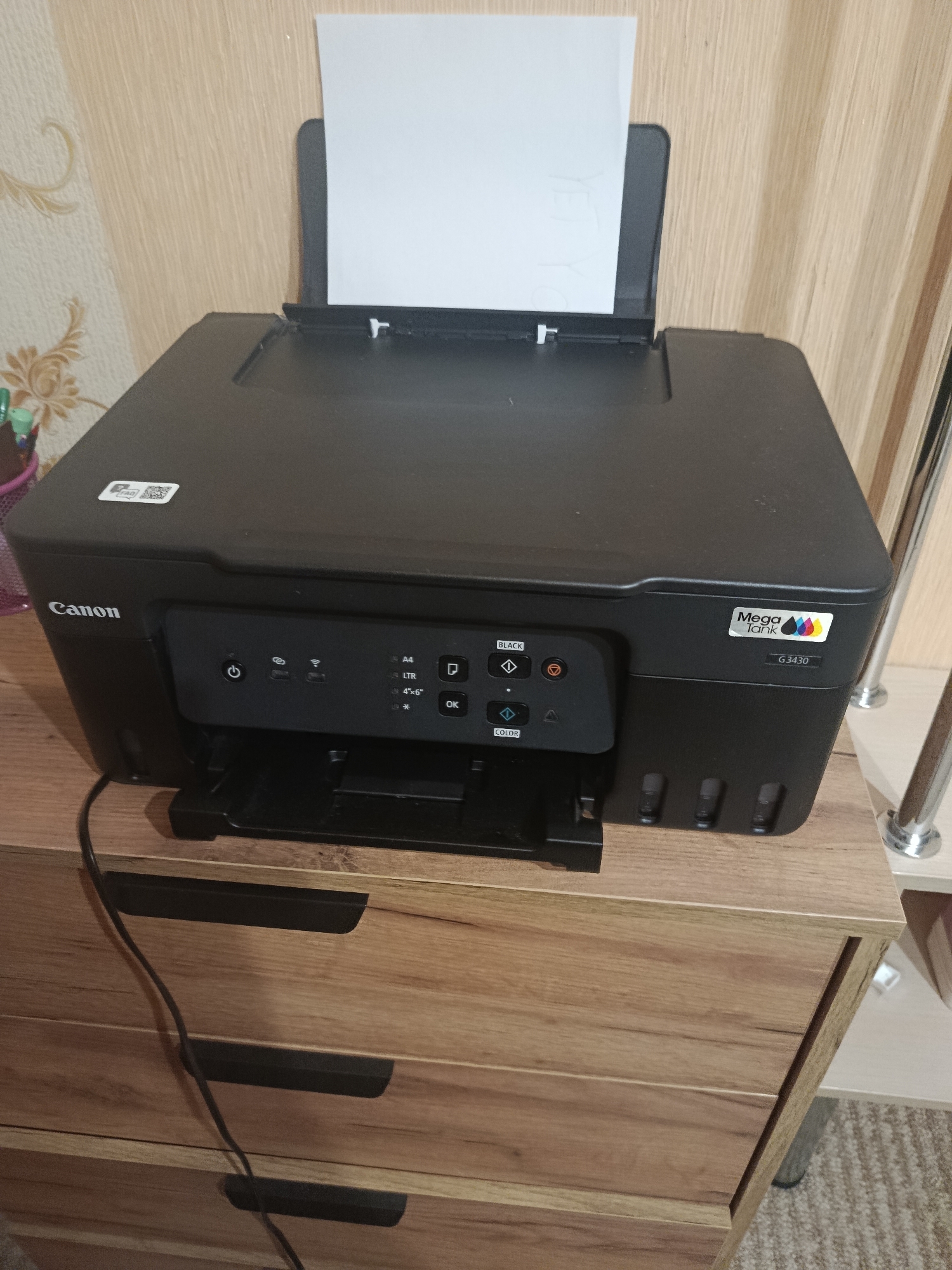 A black Canon Mega Tank G3430 printer is positioned on top of a light brown wooden dresser with three drawers. The printer has a paper tray at the top with a sheet of white paper partially inserted, and a paper output tray at the front. The control panel on the printer is visible, with buttons and indicator lights. The printer is connected to a black power cord that drapes down the side of the dresser. The dresser is situated on a carpeted floor. The background consists of a wood-paneled wall with a floral wallpaper pattern. The scene is indoors.