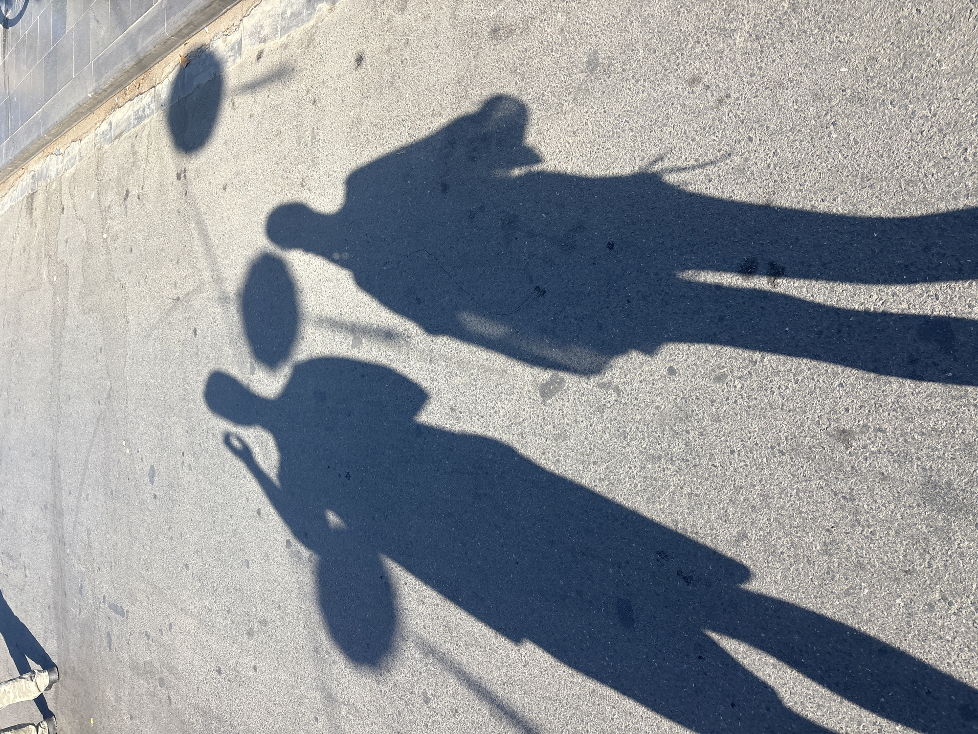 Two shadows of people are cast on an asphalt surface. The person on the left is taller, with their shadow extending longer across the ground. They appear to be holding something in their raised hand, casting a circular shadow on the ground. The person on the right is shorter, with their shadow also on the asphalt. The surface shows some wear and tear, with small dark spots and cracks visible. In the background, a concrete curb and sidewalk are visible, with some tiles laid out. The scene suggests a sunny day, with the shadows clearly defined. The specific location is Hội An Ward, Vietnam.