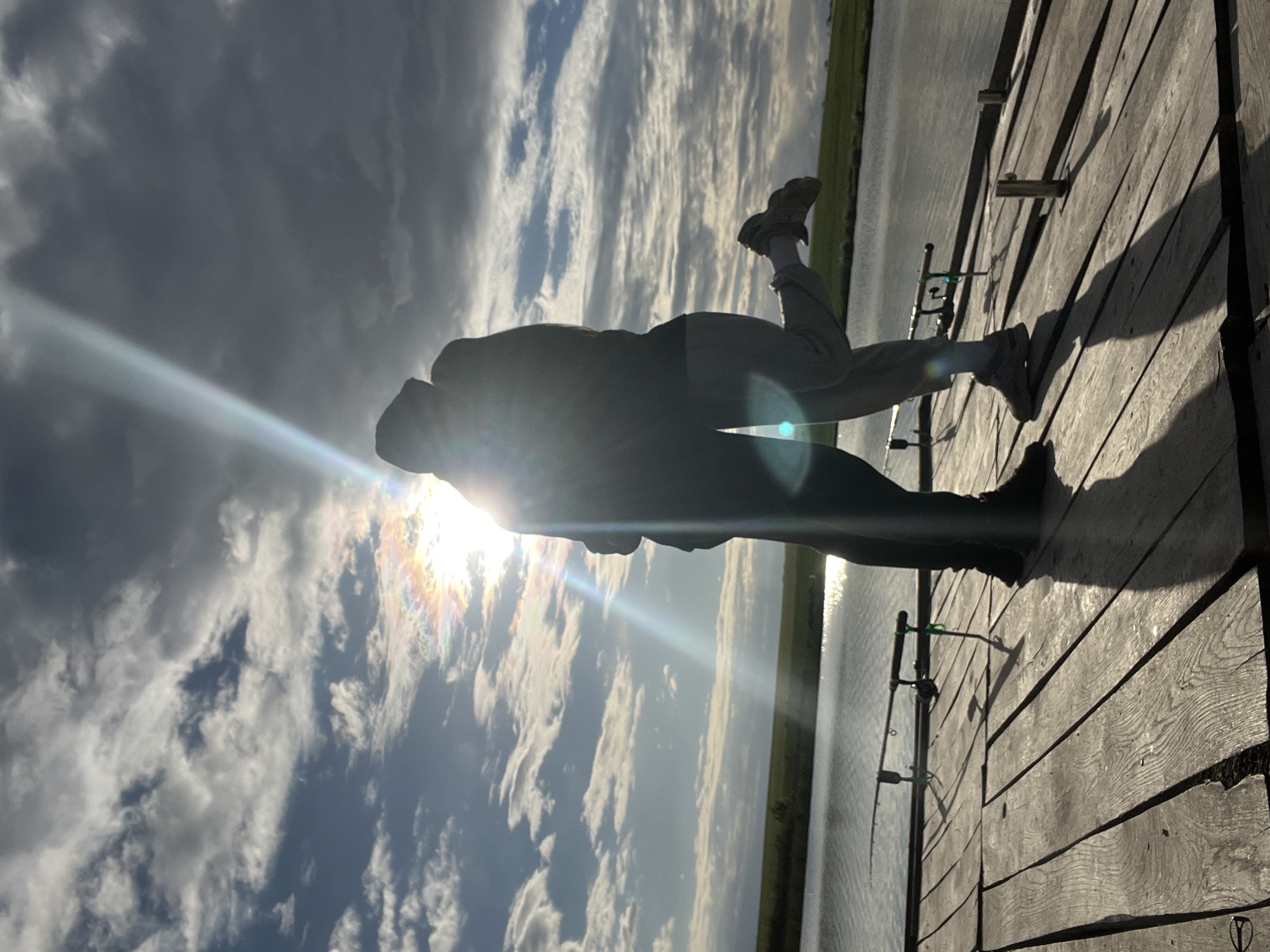 A silhouetted figure stands on a wooden structure next to a body of water, likely a lake or wide river. Two fishing rods are placed vertically, attached to the side of the wooden structure. The sky is filled with clouds, and the sun shines brightly, creating a lens flare. The location is inferred to be near Rudky, Ukraine, based on provided information.